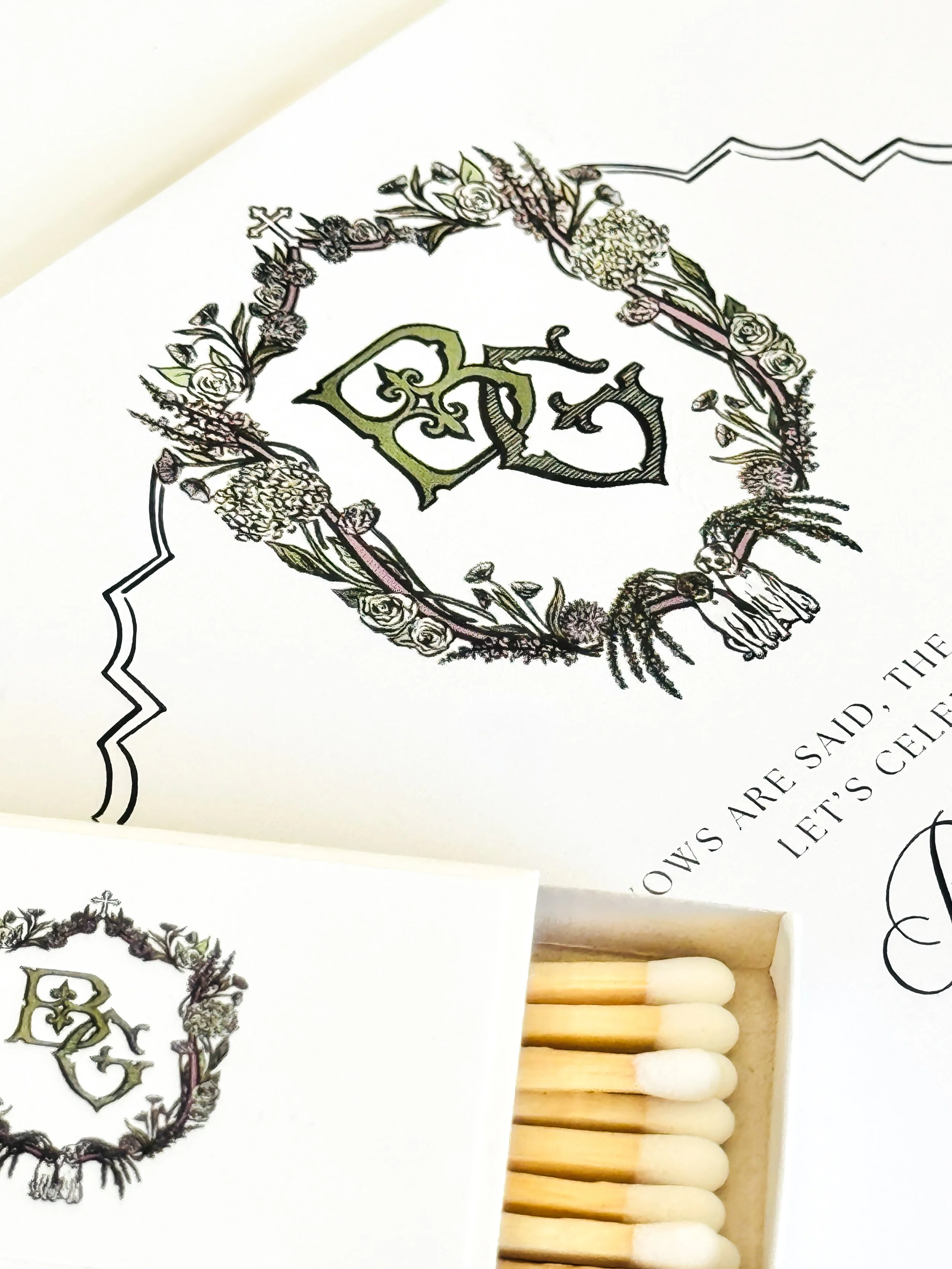 Close-up of a wedding invitation with floral wreath and stylized letters, and partially visible text underneath.