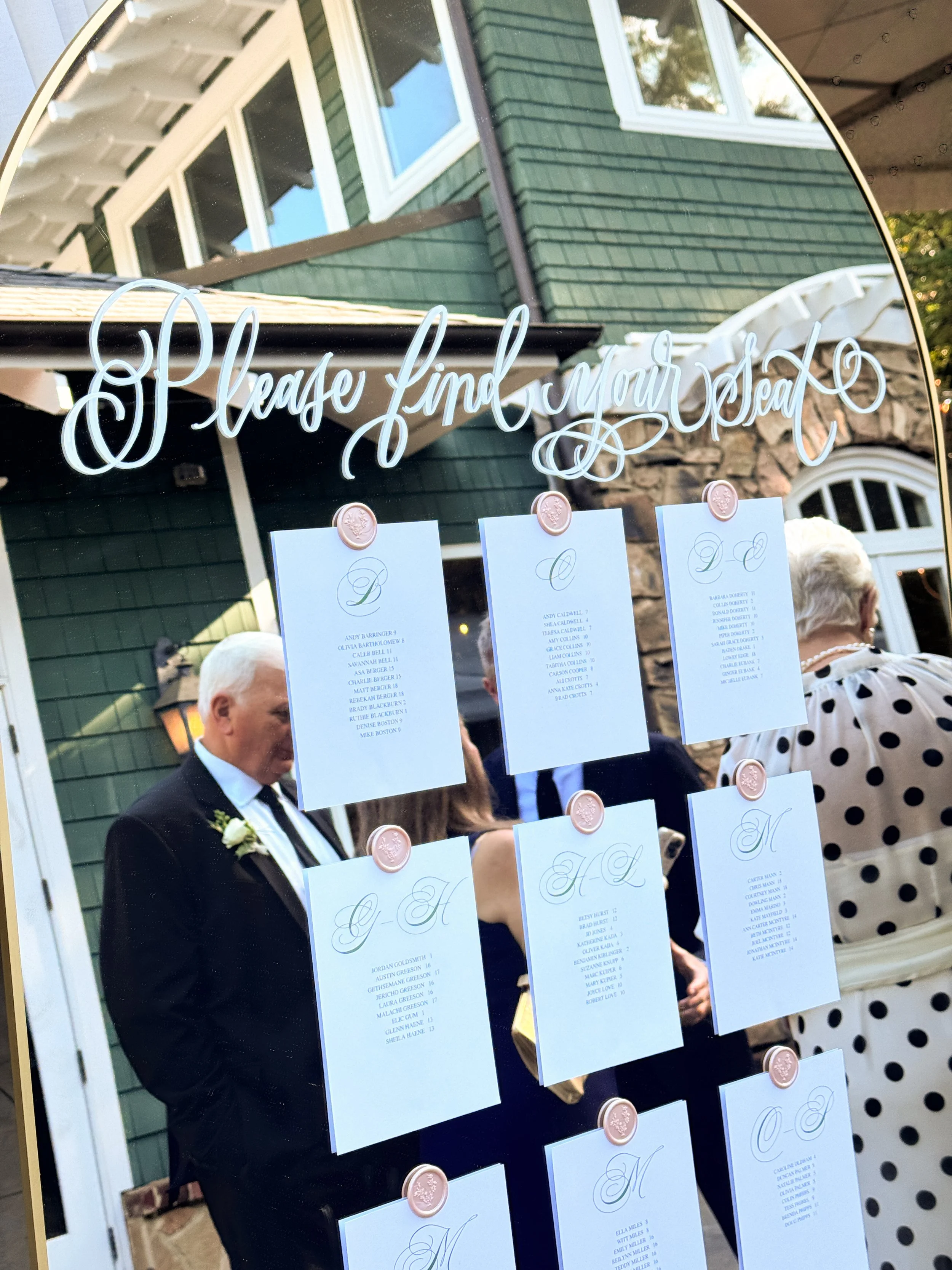 A mirror with the words 'Please Find Your Seat' written in cursive across the top, reflecting people in formal attire and a green house with white trim and multiple windows in the background.