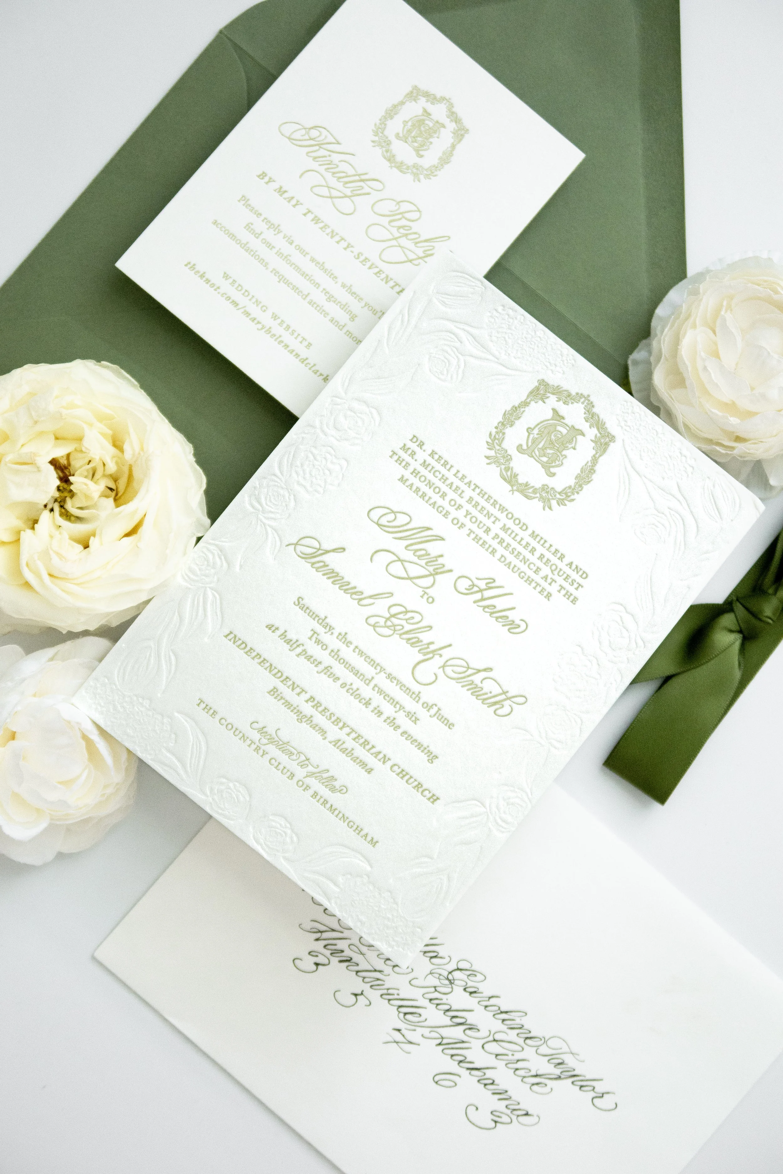 Wedding invitation cards with embossed floral and emblem designs, placed on a white surface with white roses around them.
