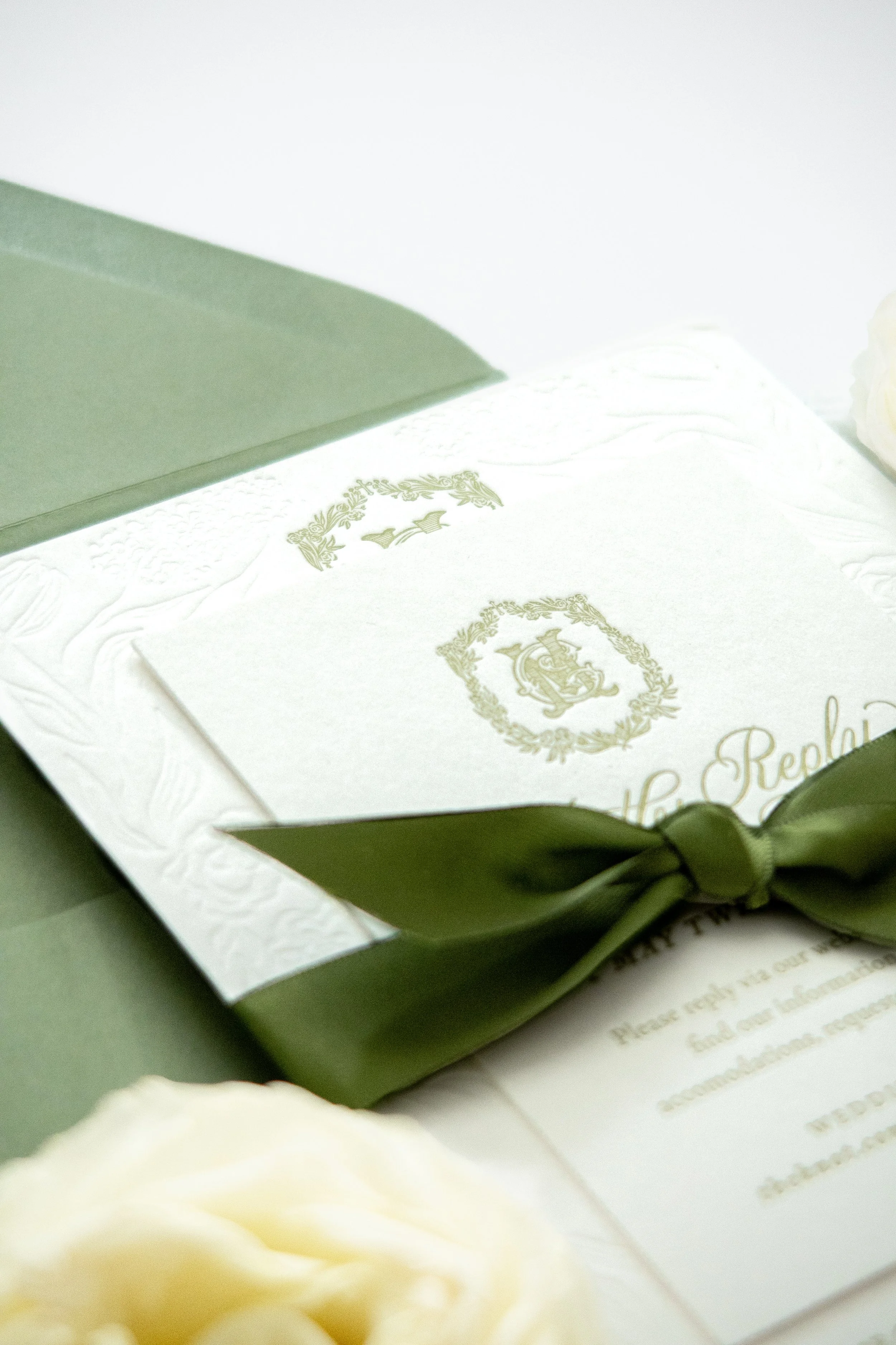A close-up of a white wedding invitation card with green ornate design and a smaller white envelope in a green box lined with white fabric, with a white flower in the foreground.