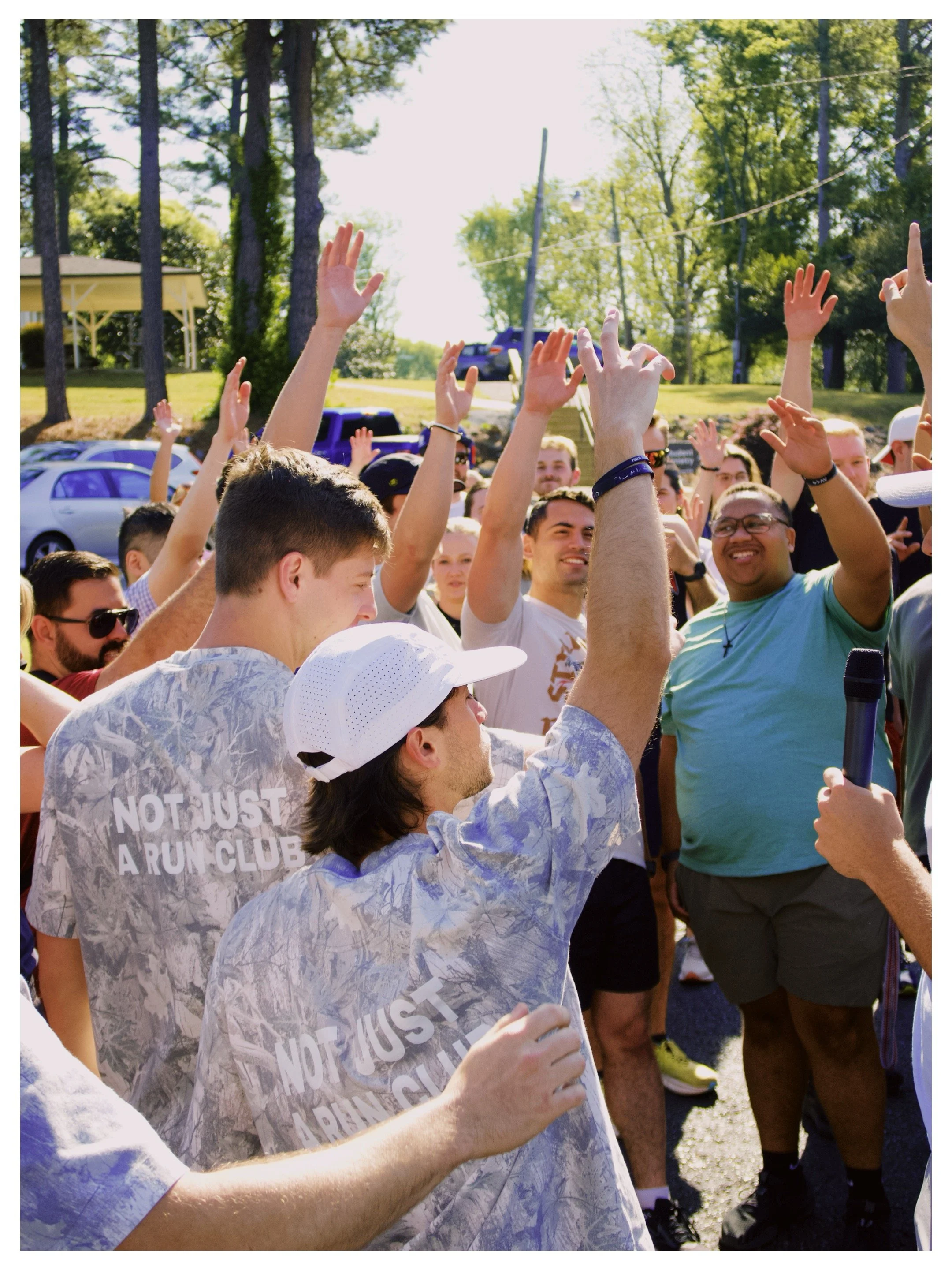 A group of people outdoors at a sunny run club, raising their hands, some smiling, some wearing sunglasses, with trees and parked cars in the background.