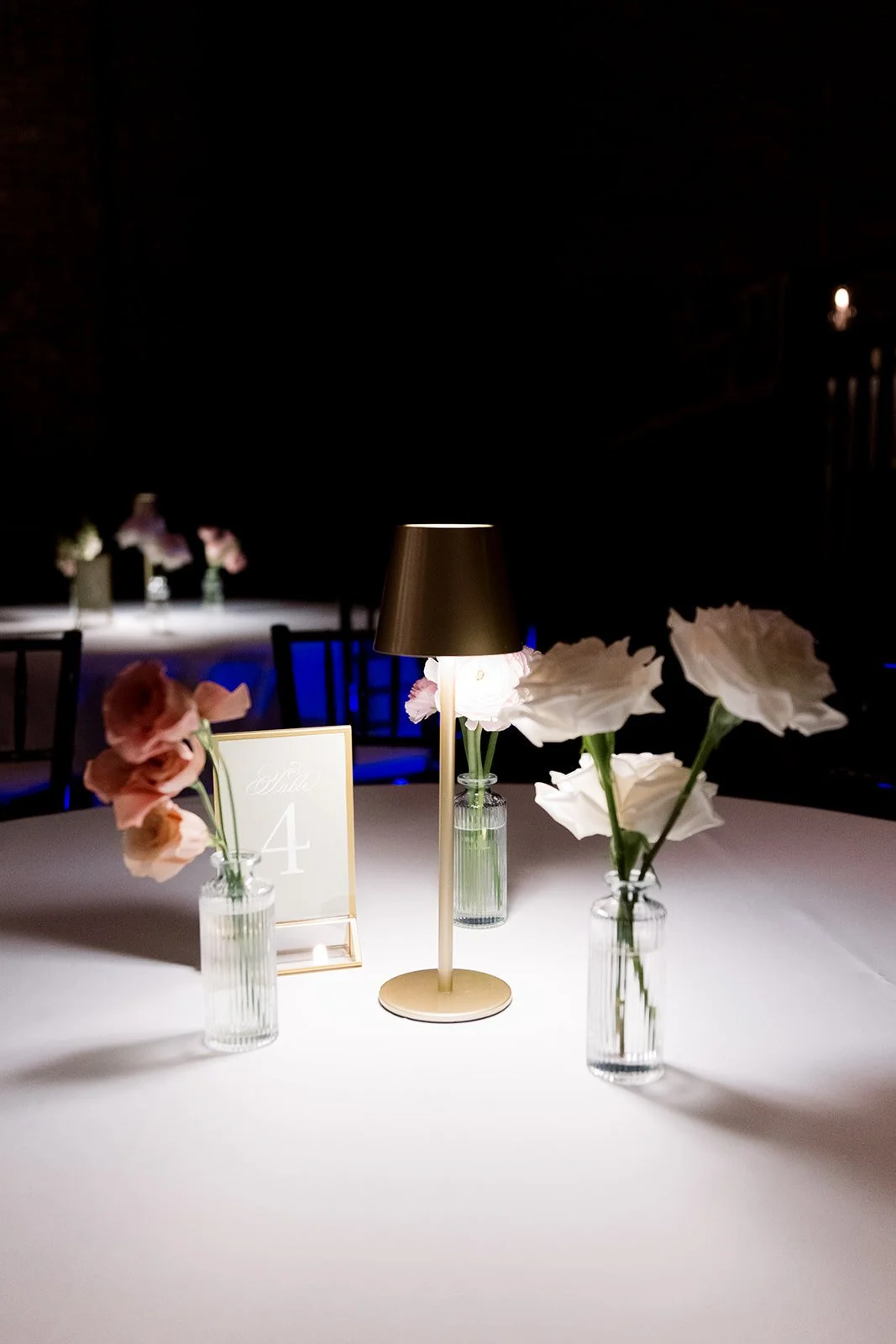 Elegant round table decorated with a gold lamp, three glass vases with white and pink flowers, and a table number sign indicating table 4, set in a dimly lit venue for a formal event.