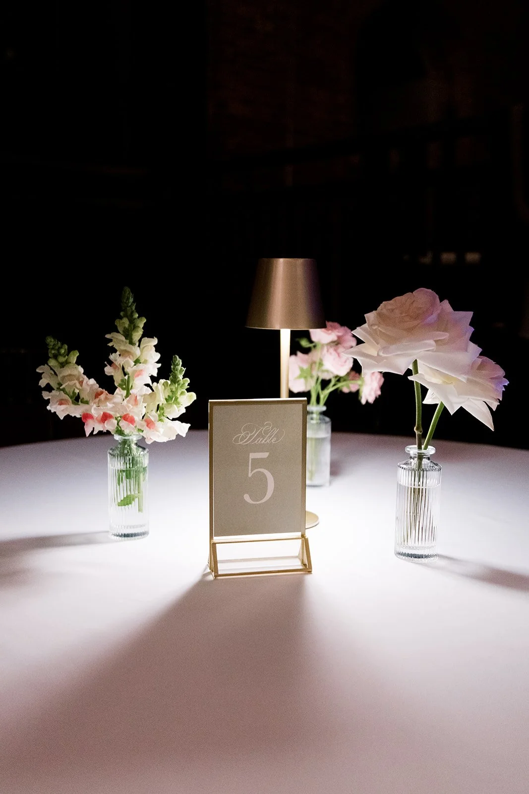 Table centerpiece with three glass vases holding pink and white flowers, a table number sign reading 'Table 5', and a small table lamp with a gold shade on a white tablecloth.