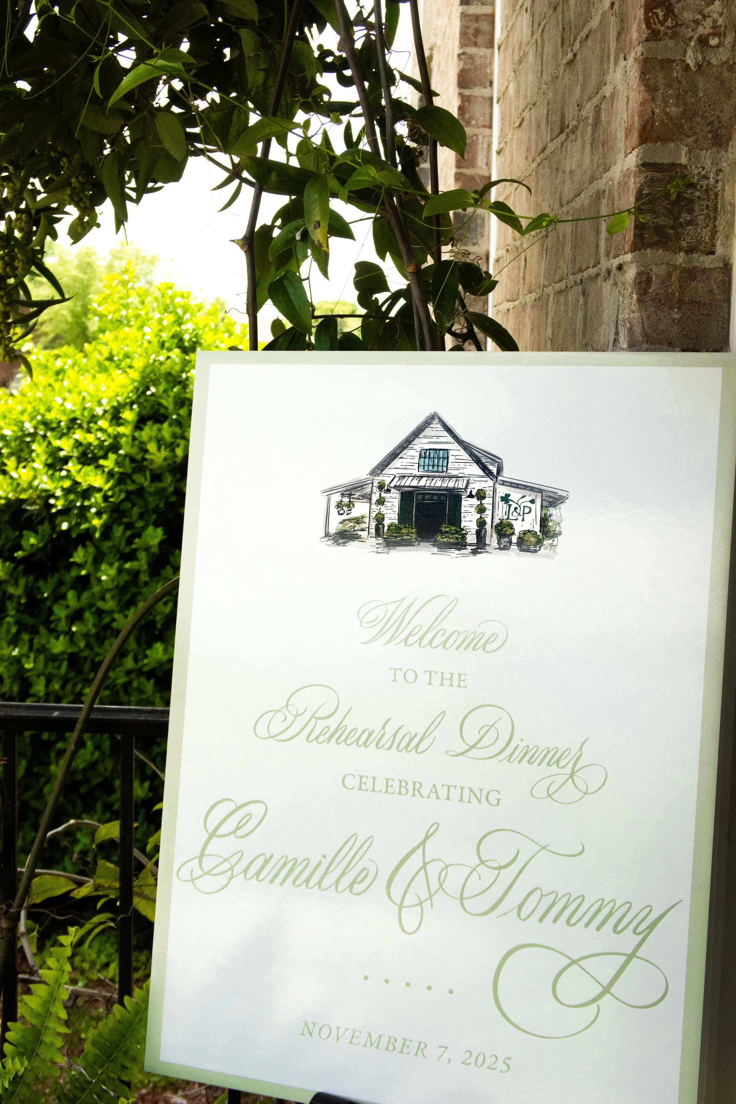 A white wedding rehearsal dinner invitation with an illustration of a house at the top, surrounded by planters and greenery, and text that reads "Welcome to the Rehearsal Dinner celebrating Camille & Tommy, November 7, 2025." The sign is outdoors near a brick wall, with bushes and trees in the background.