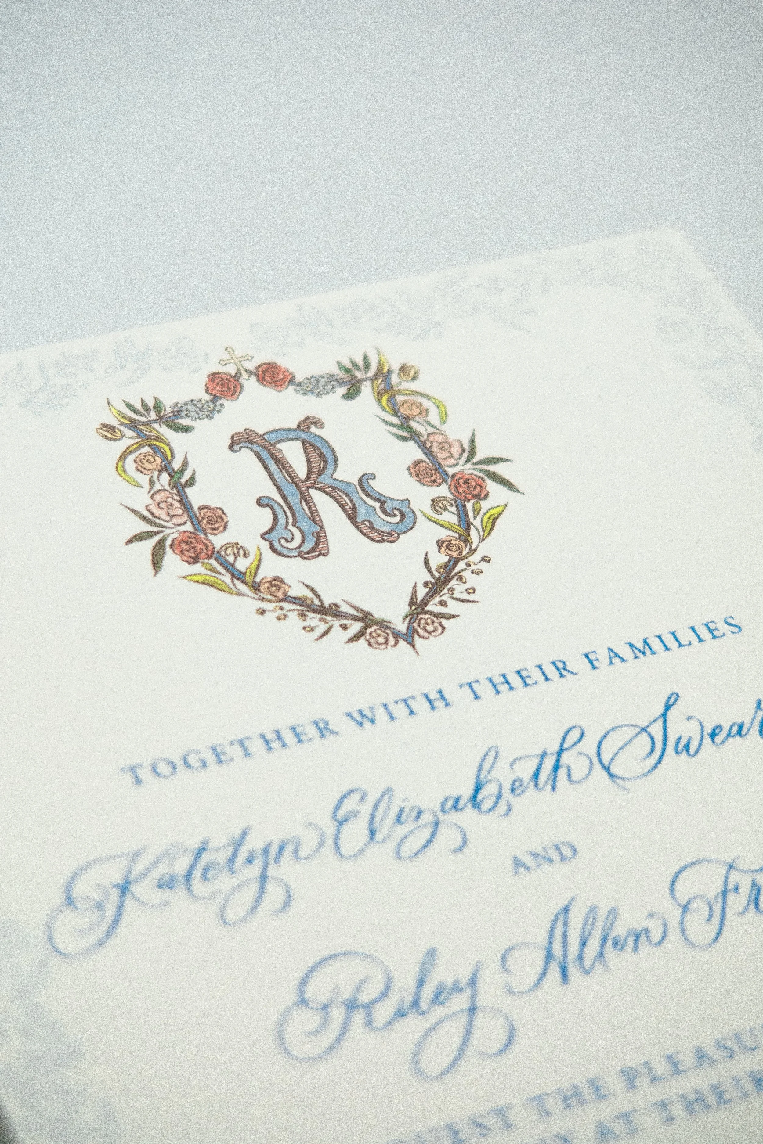 A close-up of a wedding invitation with a decorative floral crest featuring the letter 'R' in the center, and a paragraph of text in blue cursive and regular font.