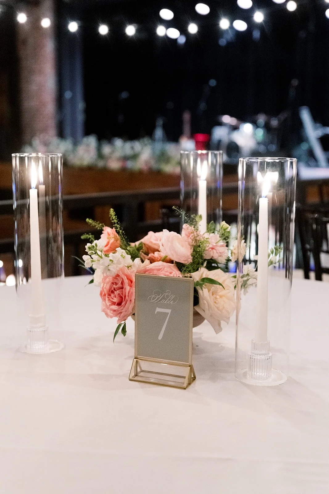 Elegant wedding table centerpiece with pink and white roses, candles in tall glass holders, and a table number sign reading 'Table 7' in a softly lit venue.