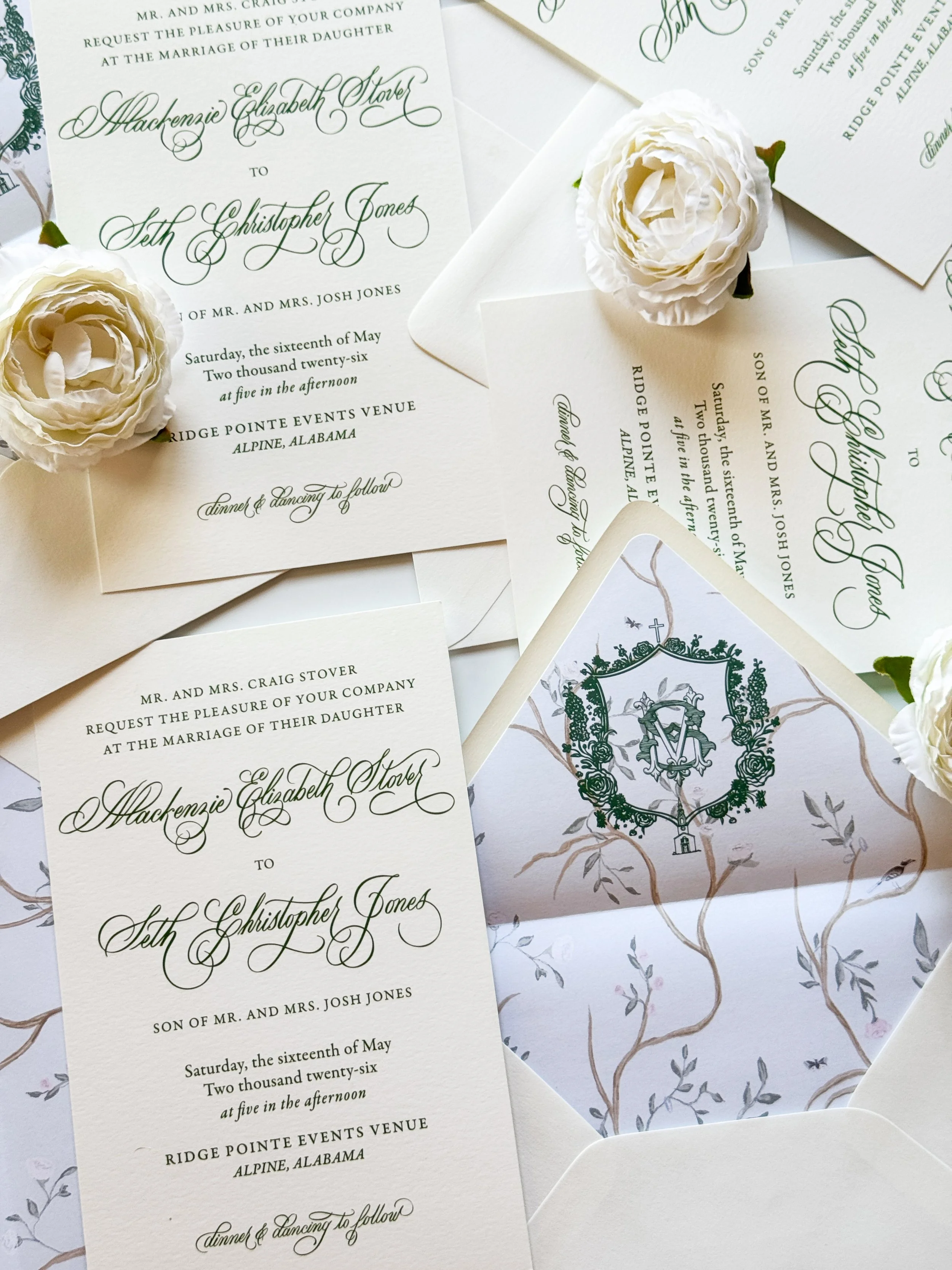 Several elegant wedding invitations with cursive green text, alongside white flowers and a decorative card with a crest, arranged on a white surface.
