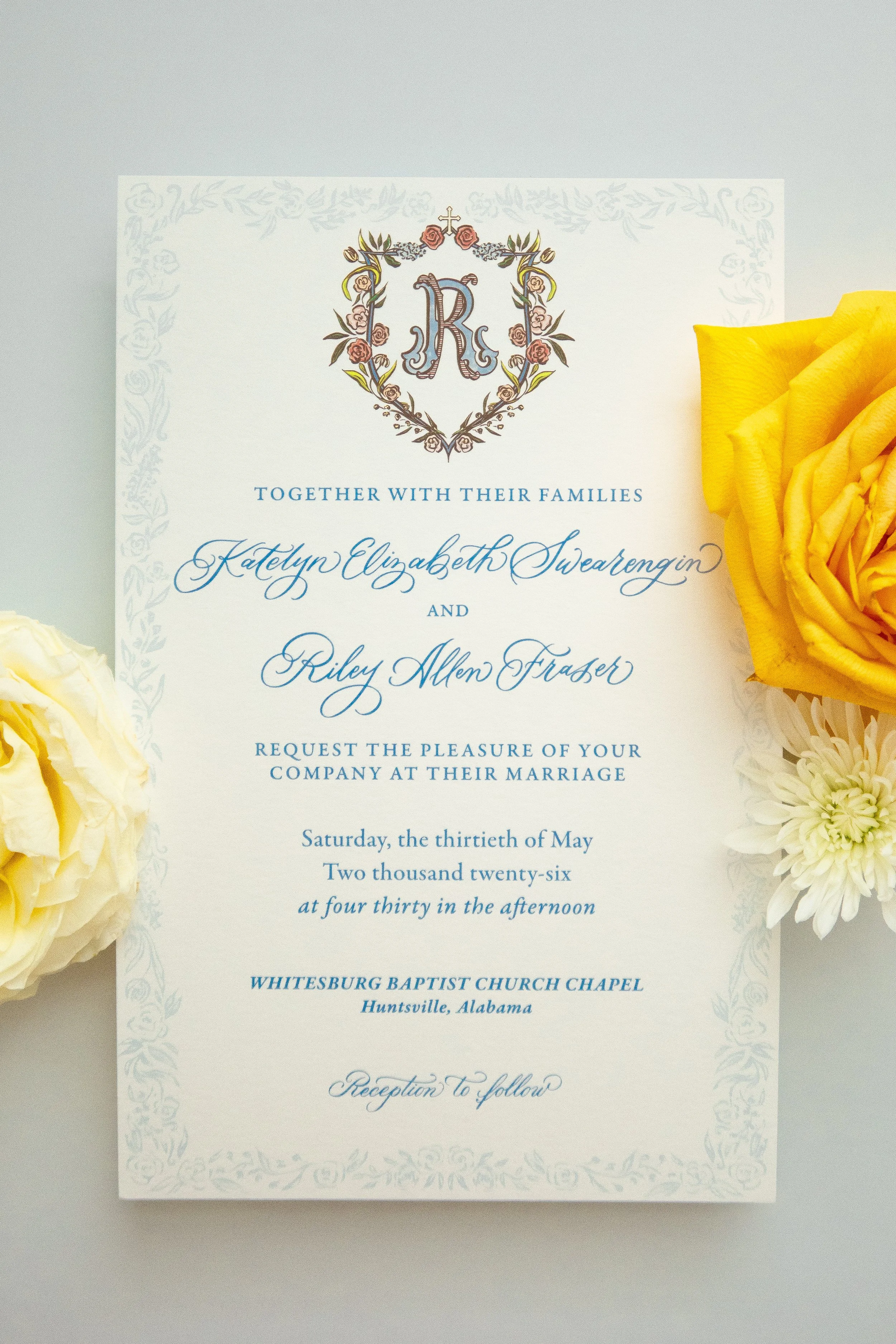 Wedding invitation card with floral border, featuring initials R and K within a crest made of florals, and yellow and white flowers beside it.