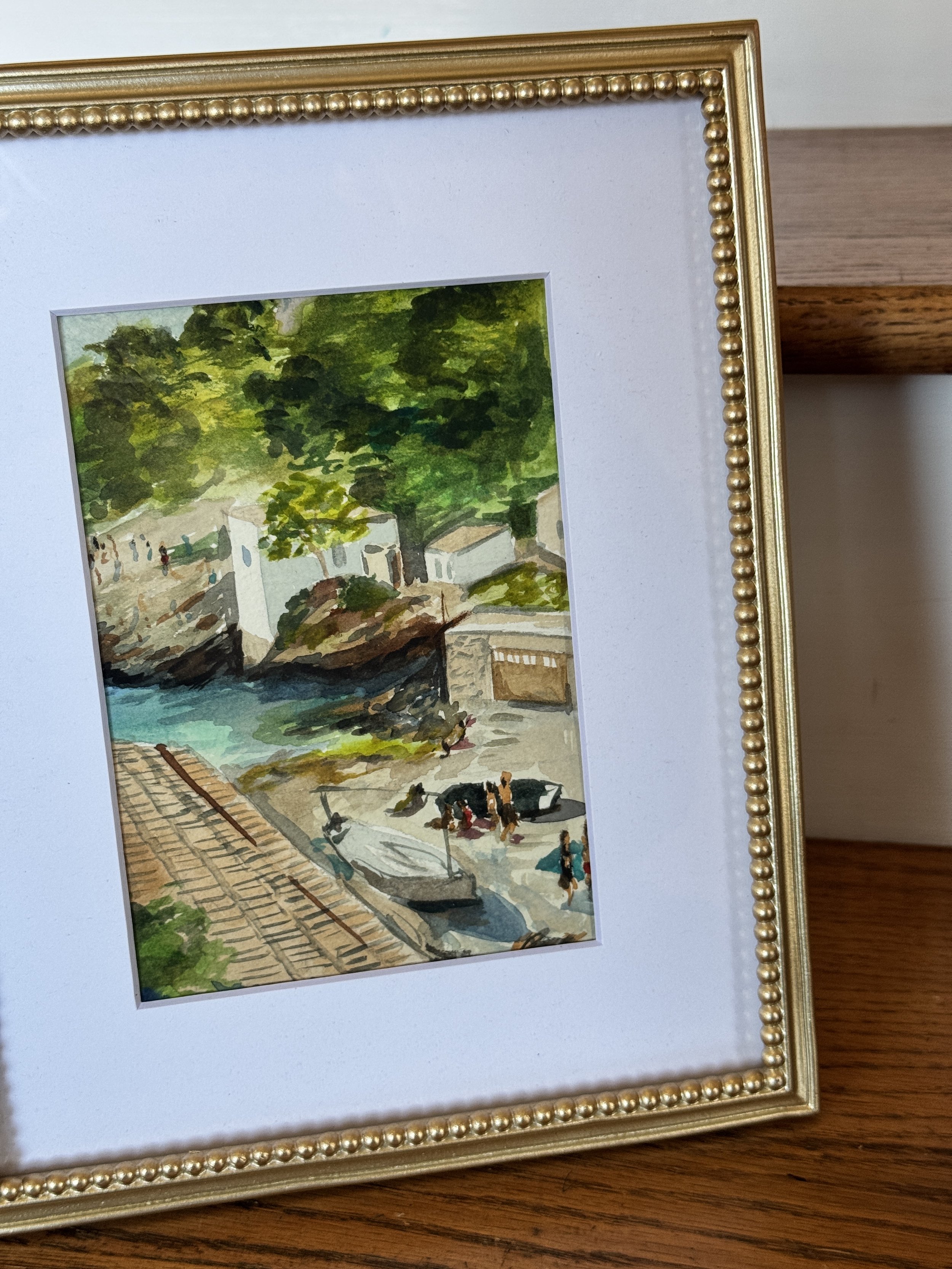 Framed watercolor painting of a lakeside scene with trees, boats, and buildings, with people walking nearby.