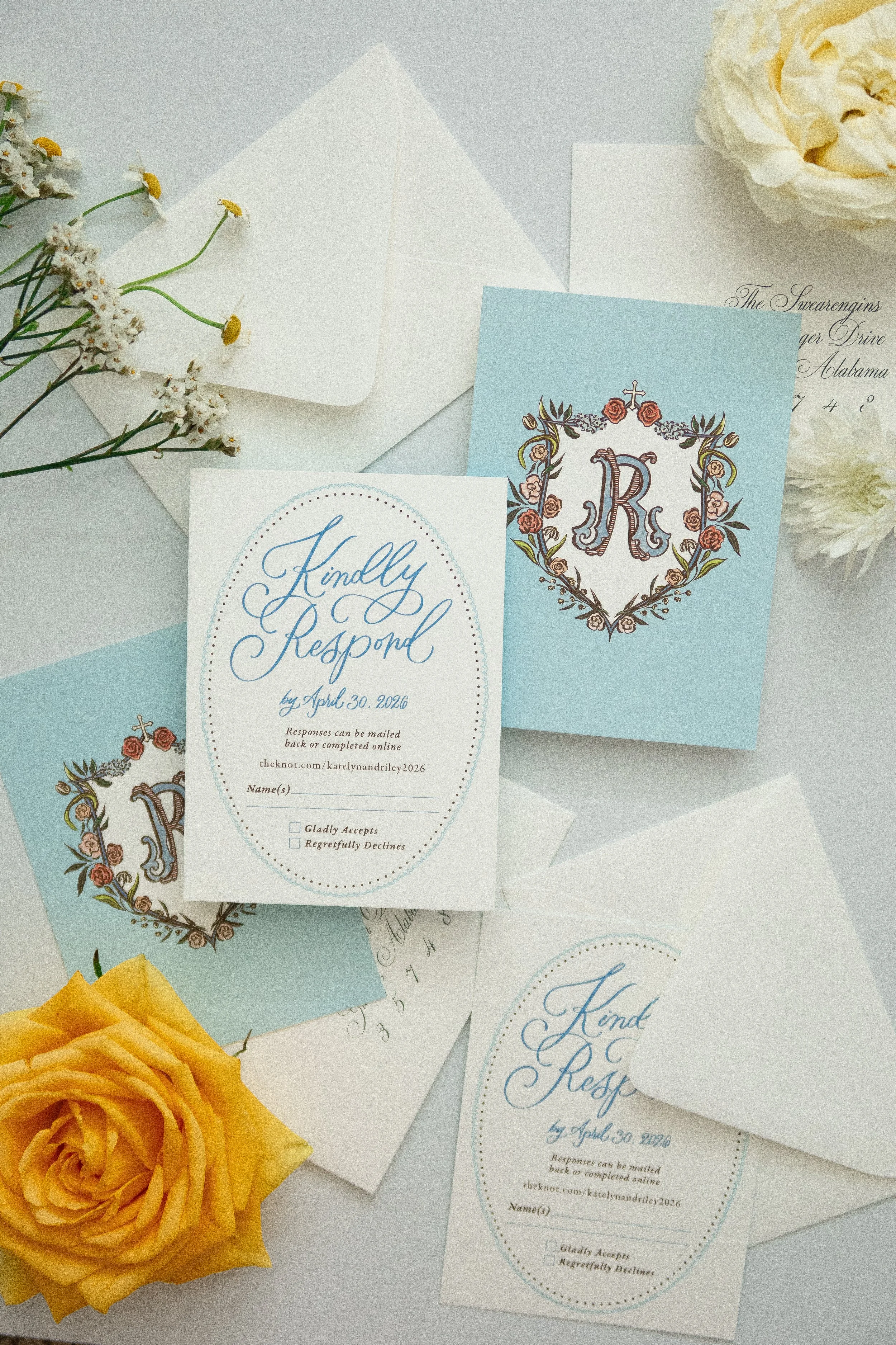 A collection of wedding response cards with a religious monogram, white and blue paper, an open white envelope, yellow roses, and small white flowers on a light surface.