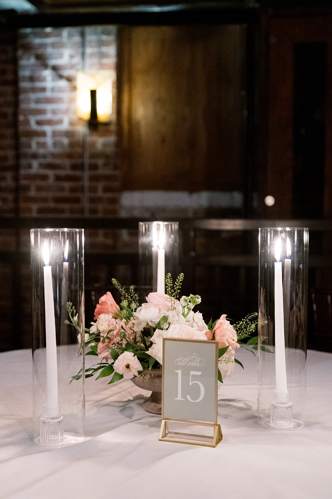 A table decorated with a floral arrangement, three tall glass candle holders with lit white tapers, a table number sign reading 'Table 15', and a white tablecloth, set against a dimly lit background with brick wall and a wall lamp.