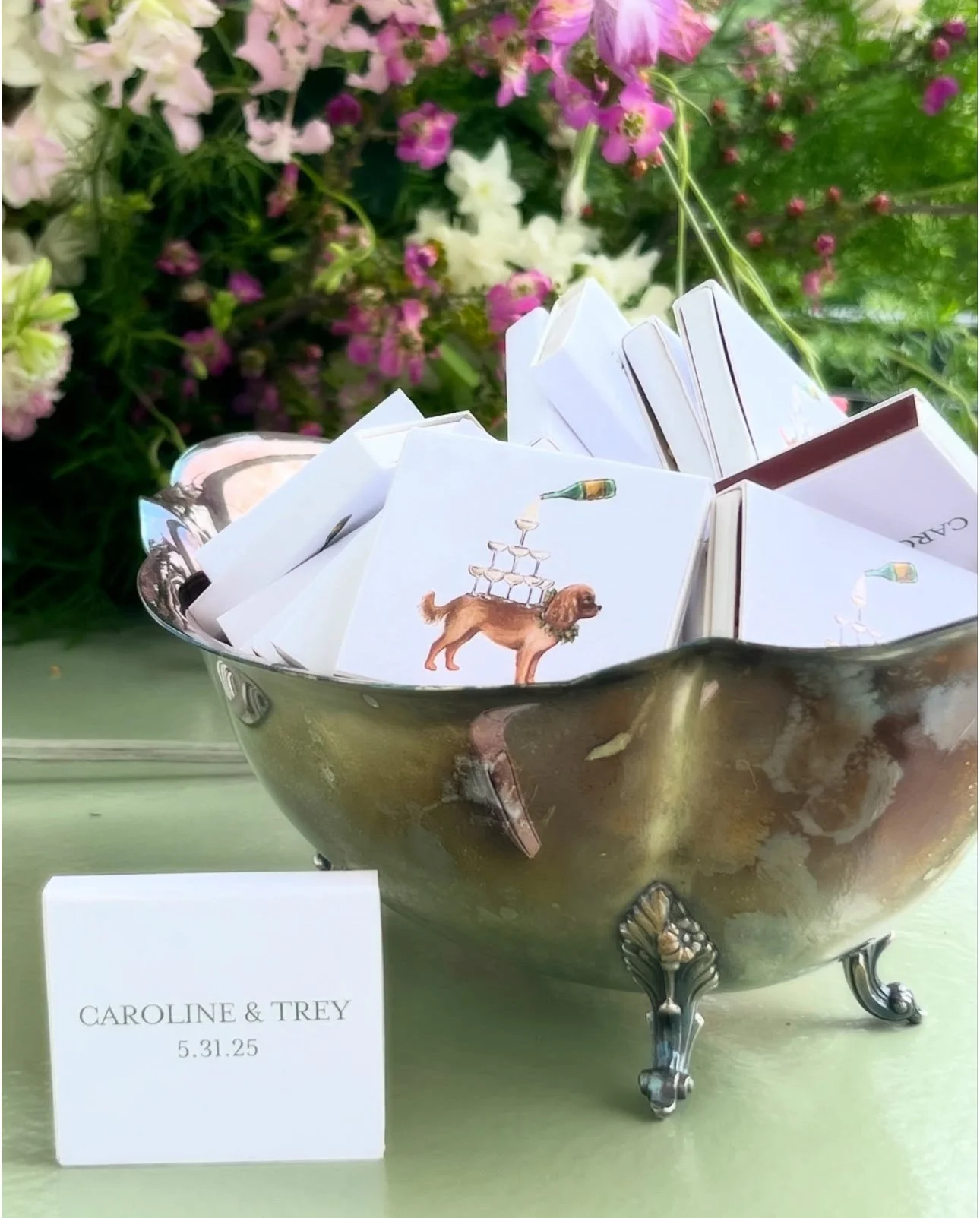 A silver decorative bowl filled with small white matchboxes, some featuring a dog illustration with stacked glasses and a bottle on its back. Pink, white, and purple flowers in the background. 