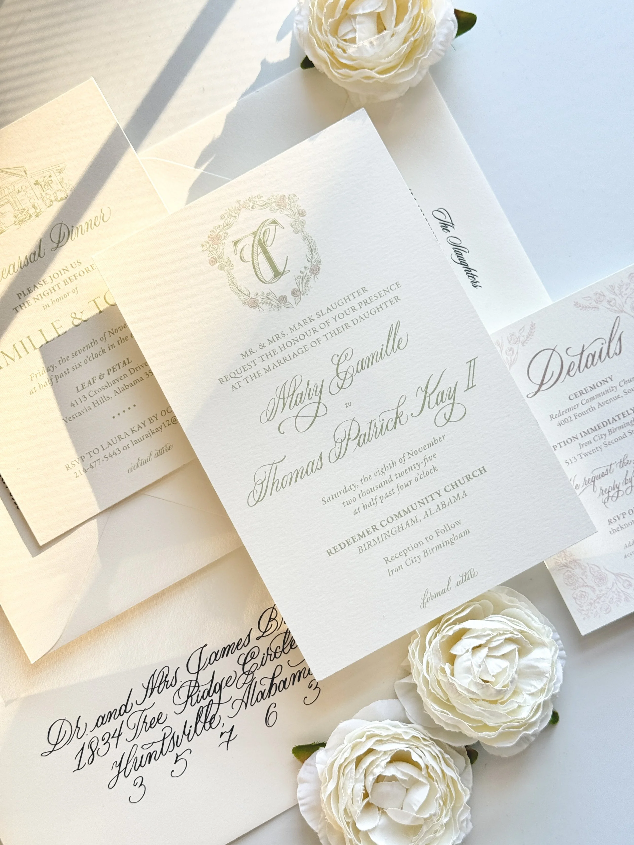 Wedding invitation cards with elegant cursive writing and decorative monogram, arranged on a white surface alongside white artificial roses.