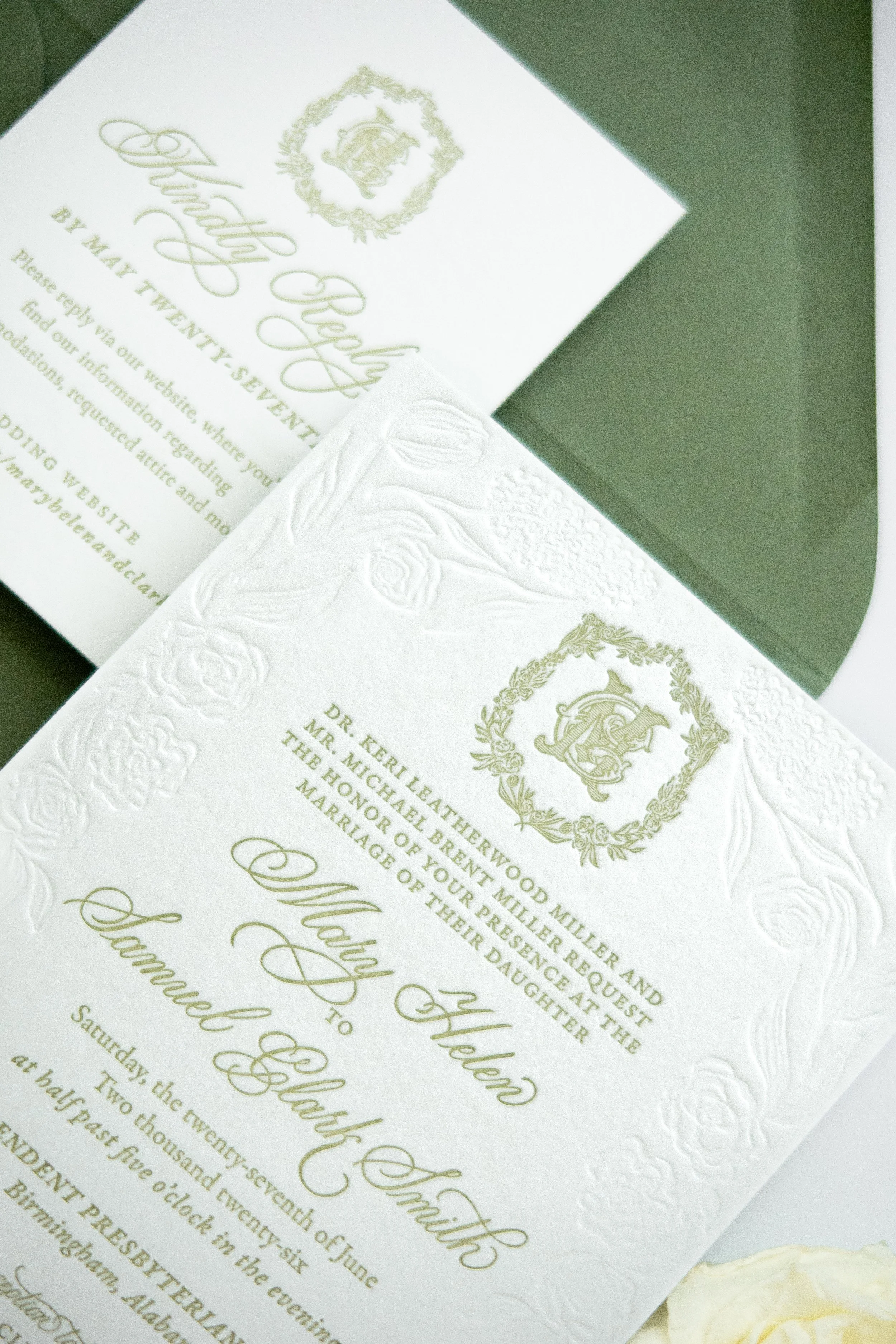 Close-up of a white wedding invitation card with green embossed text and a floral embossed border, accompanied by a white rose.
