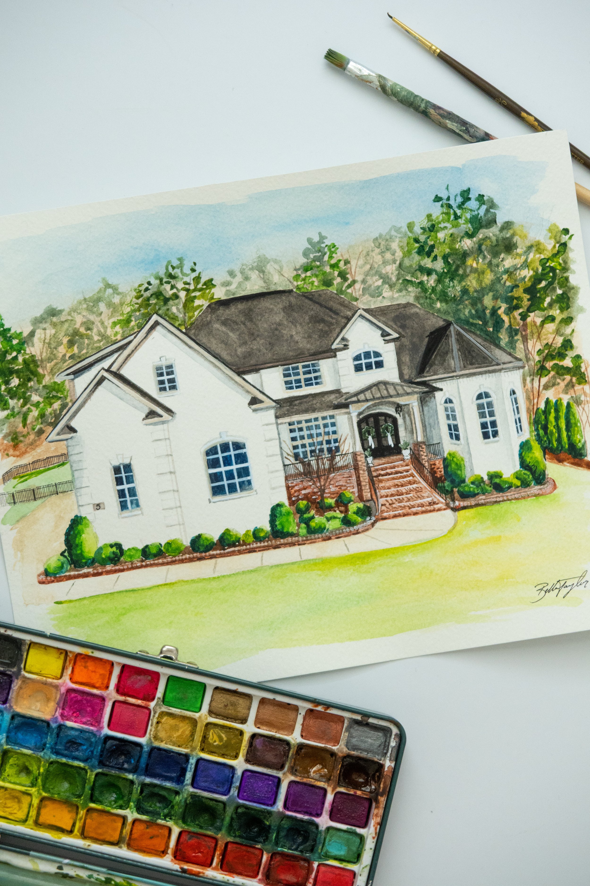 Watercolor painting of a two-story white house with dark roof, front steps, and landscaped front yard, with watercolor paints and brushes nearby.