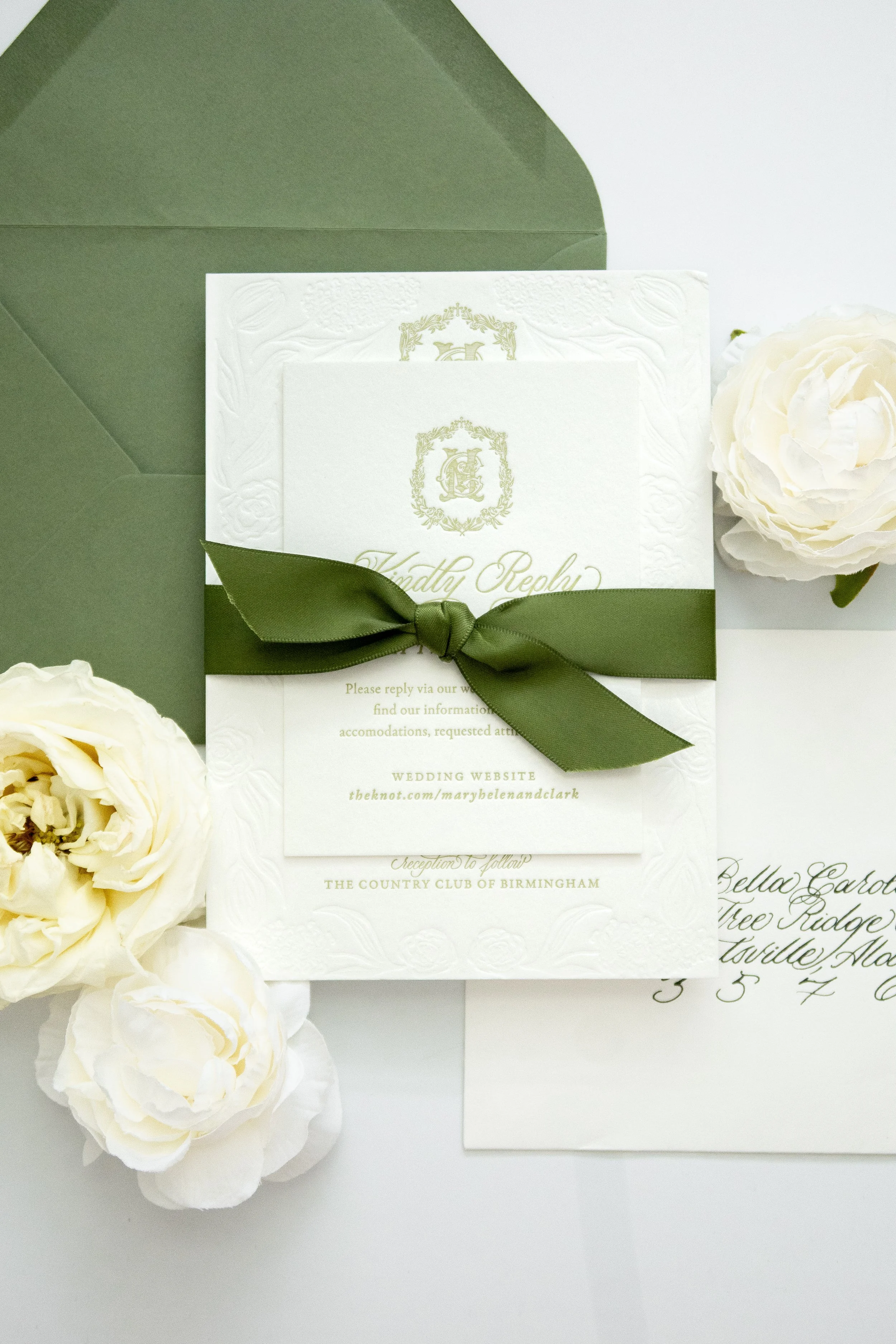 A wedding invitation set by Bella Taylor with white textured cards, decorated with gold crest and text, tied with a dark green ribbon. Surrounding it are white roses and a handwritten letter.