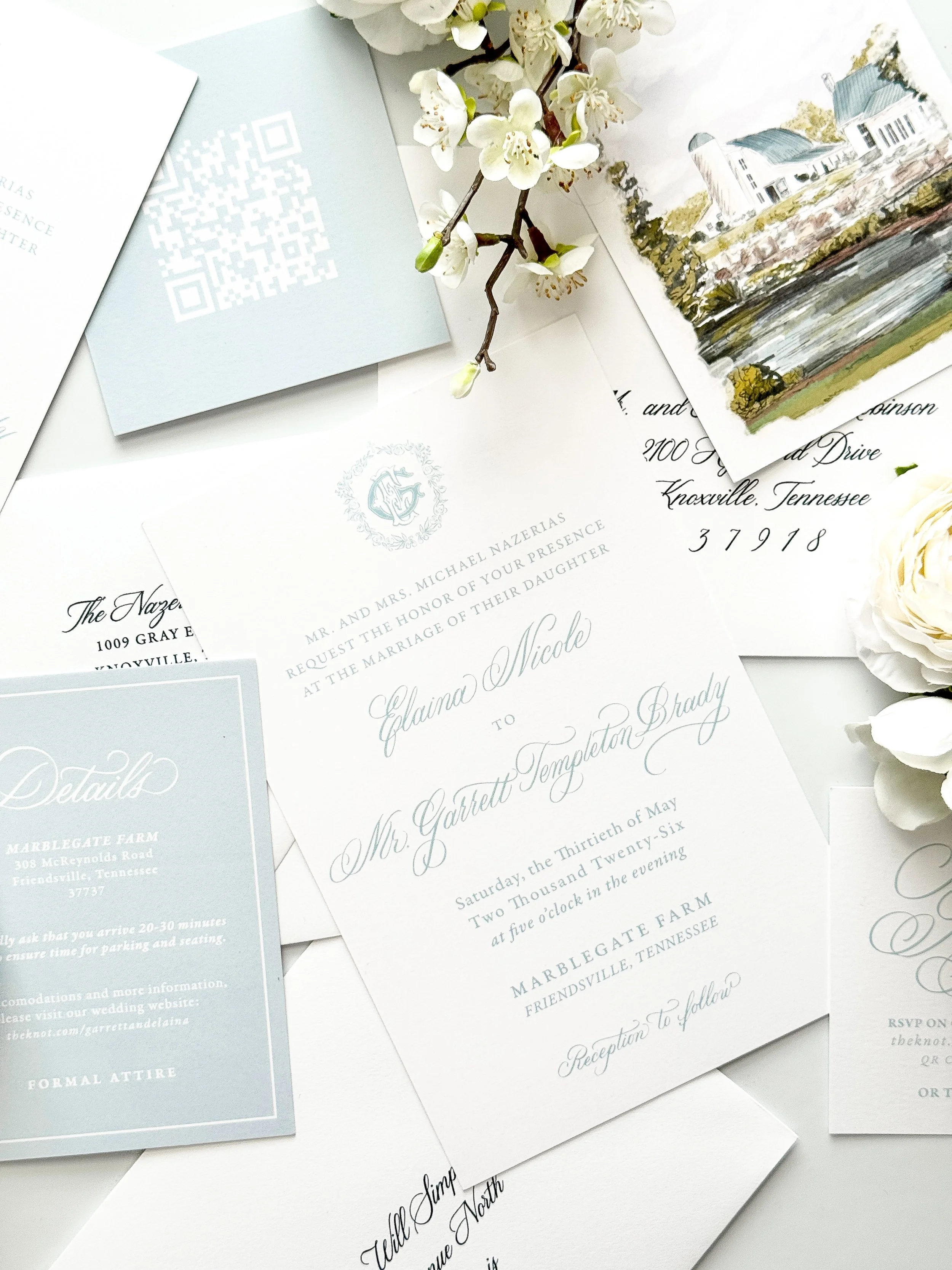 An assortment of wedding invitation cards, a branch with white flowers, and a hand-painted picture of a building in the background.