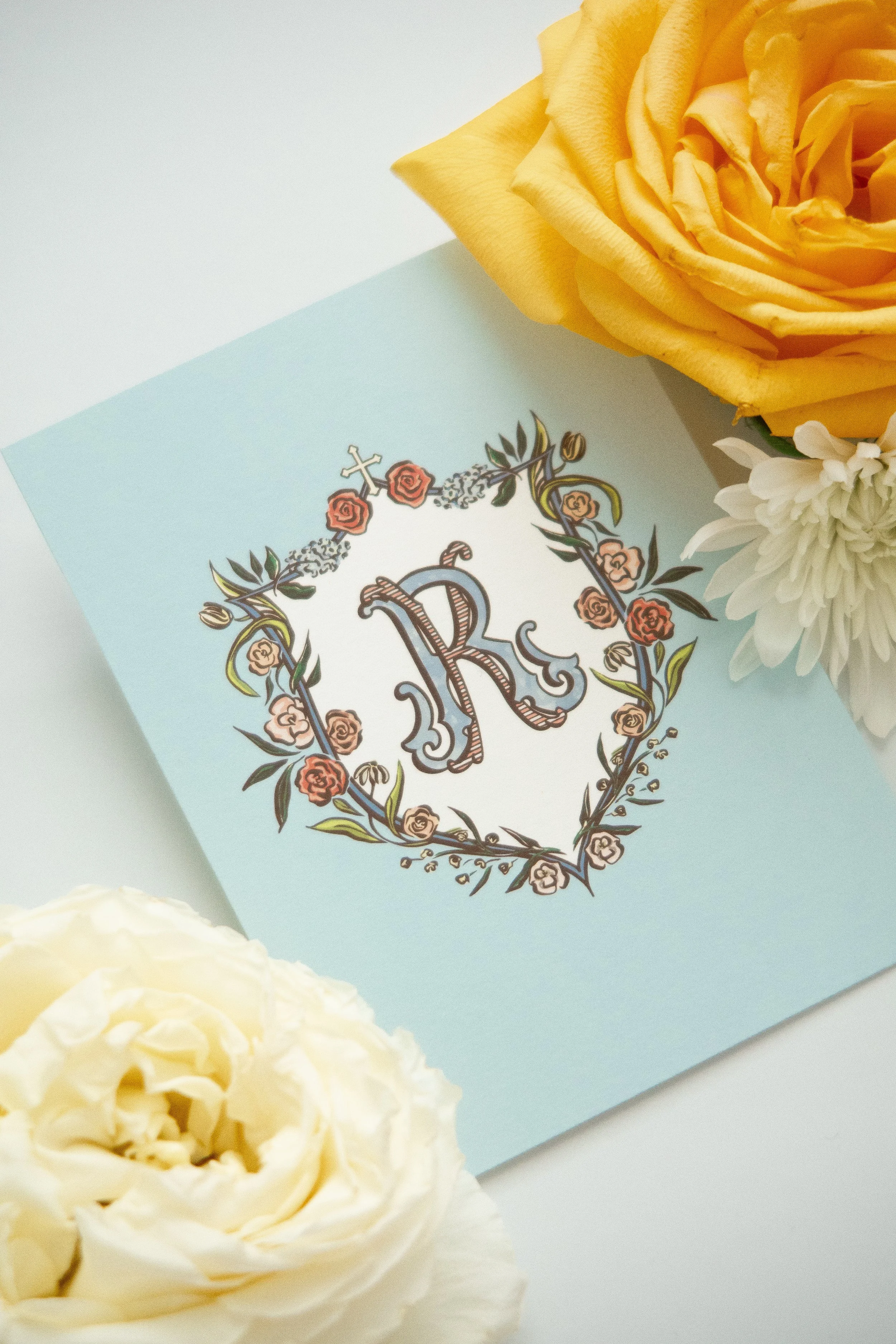 A light blue card with a decorative border of roses and intricate patterns, with linked Christian crosses, placed next to a white flower and a large yellow rose.