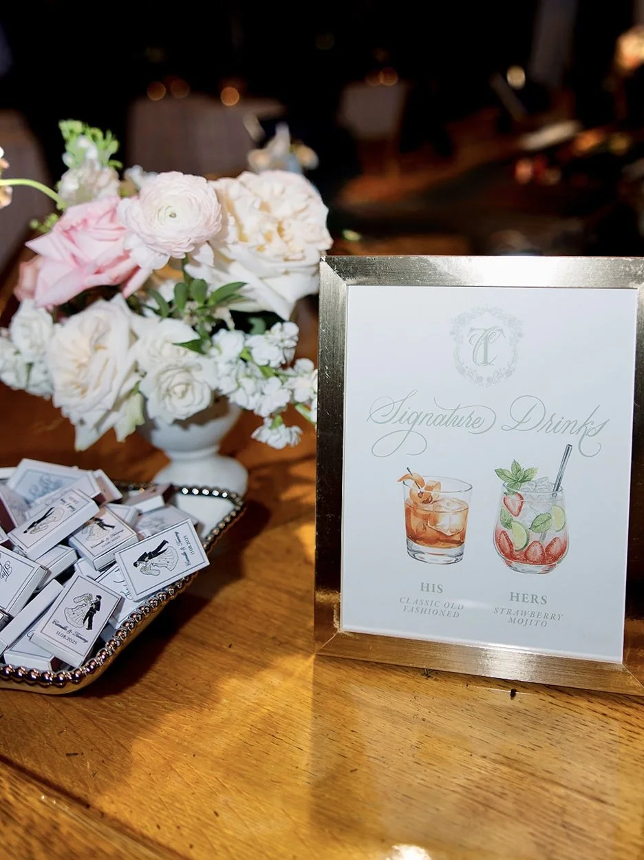 A table decorated with a pink and white flower arrangement in a white vase, a silver tray with matchboxes, and a framed sign listing signature drinks with illustrations of a classic old-fashioned and a strawberry mojito, labeled 'His' and 'Hers'.