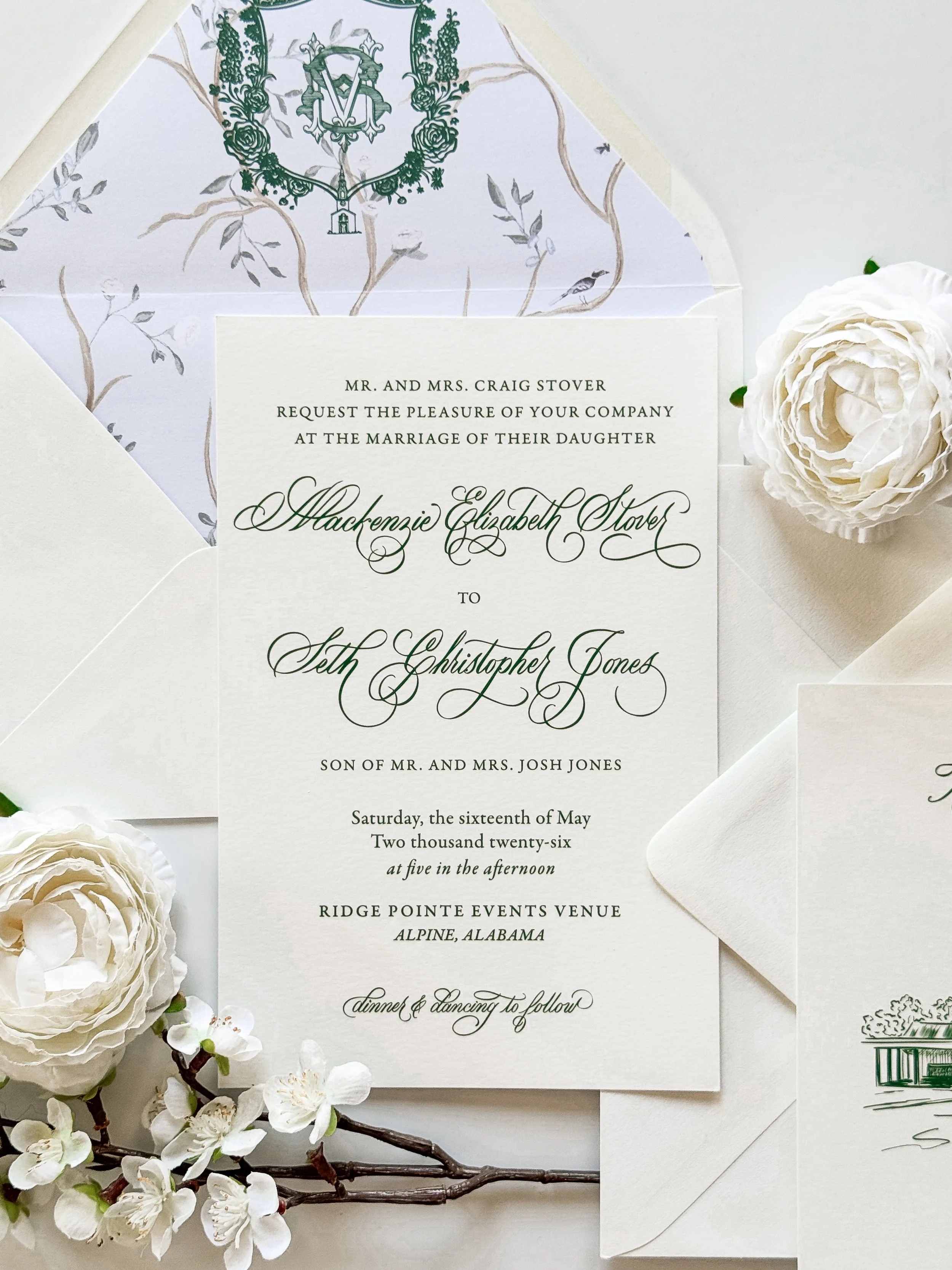 Wedding invitation card with elegant cursive font, surrounded by white flowers and a branch of blossoms, with a white envelope nearby.