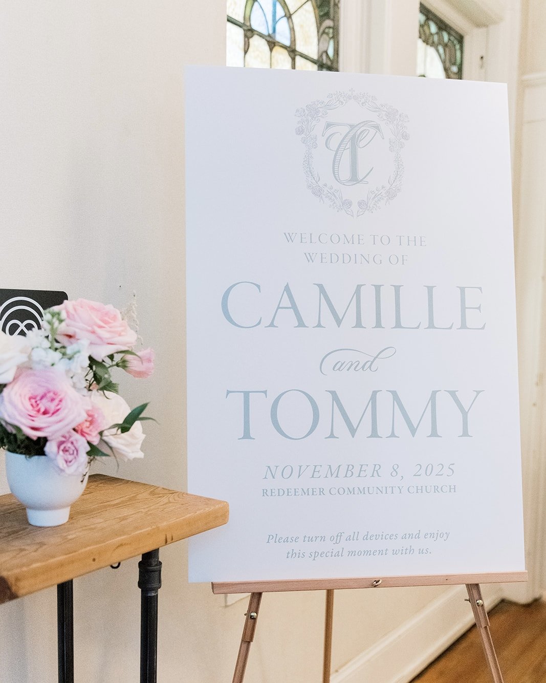 Wedding reception sign welcoming Camille and Tommy on November 8, 2025, at Redeemer Community Church, with a floral arrangement of pink and white roses on a wooden table.