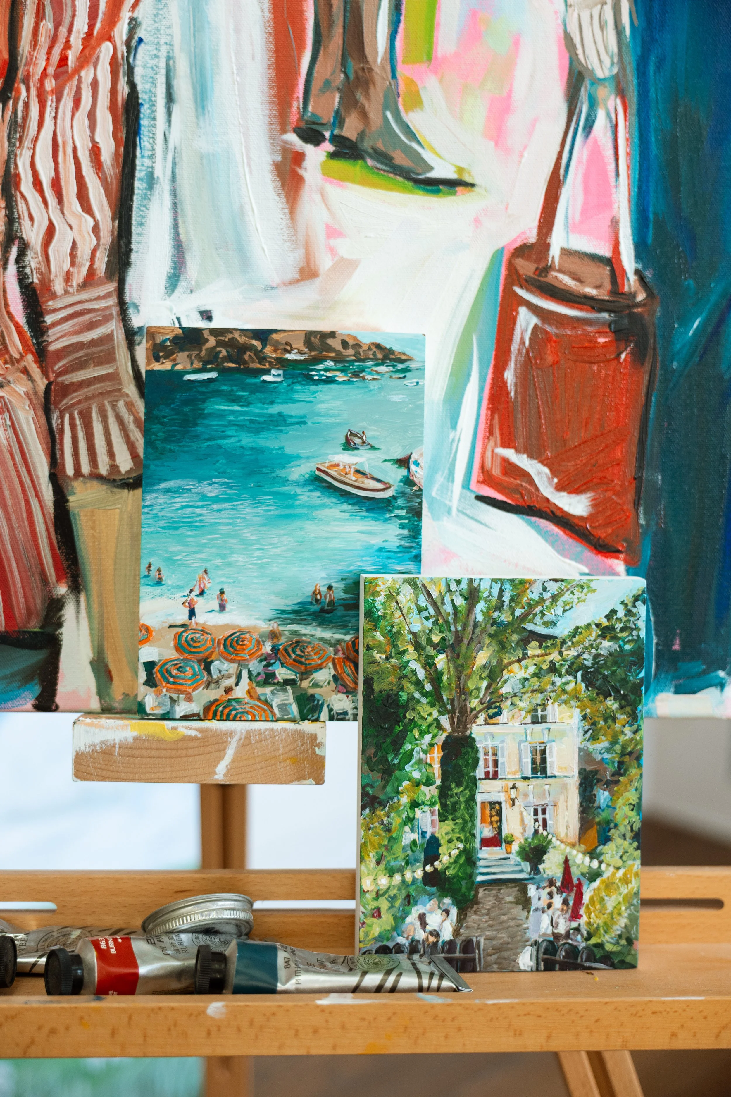 Paintings of a beach scene, a tree, and a city street scene, along with art supplies on a wooden table.