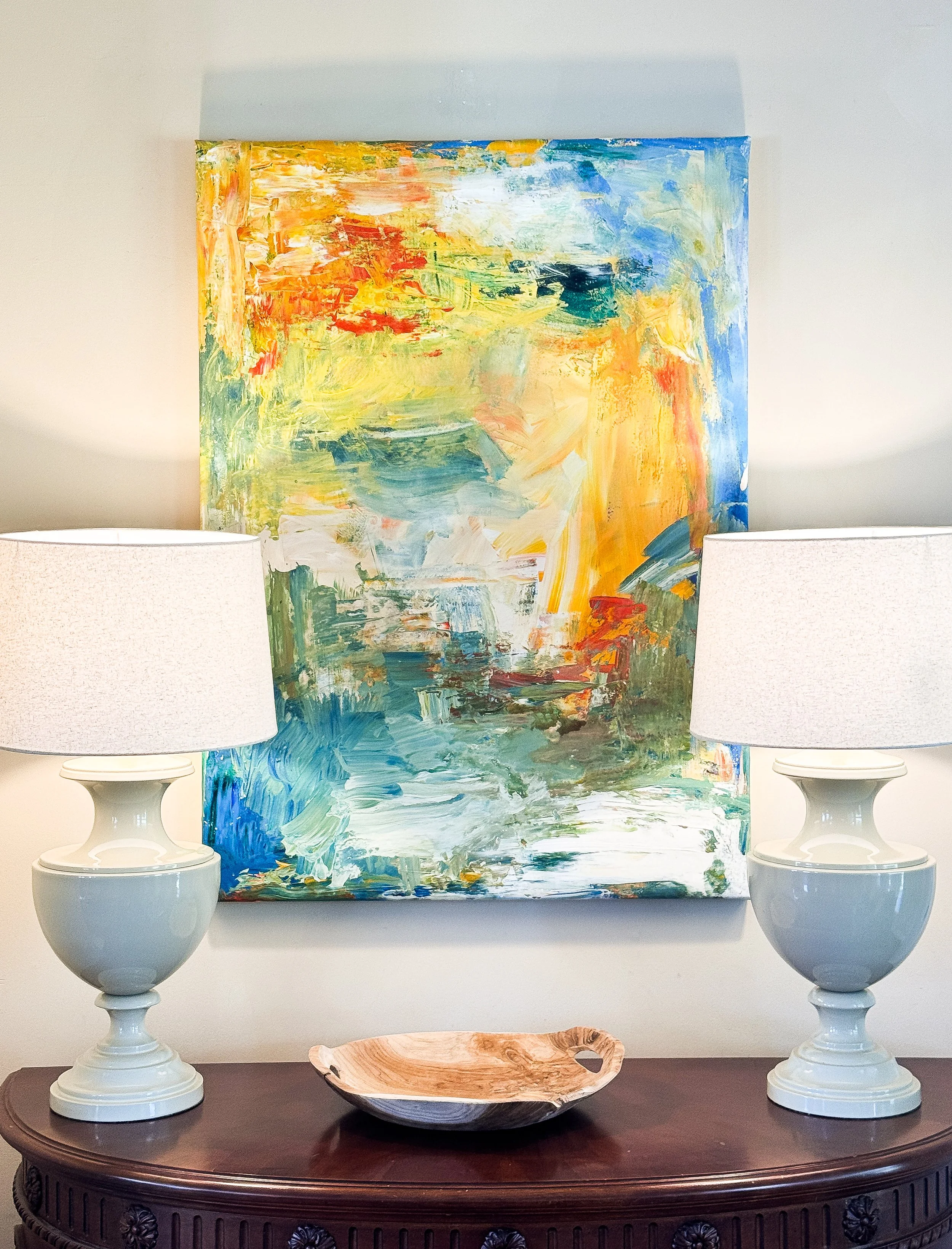 A colorful abstract painting on a wall with two white table lamps on either side and a wooden bowl on a dark wooden table in front.