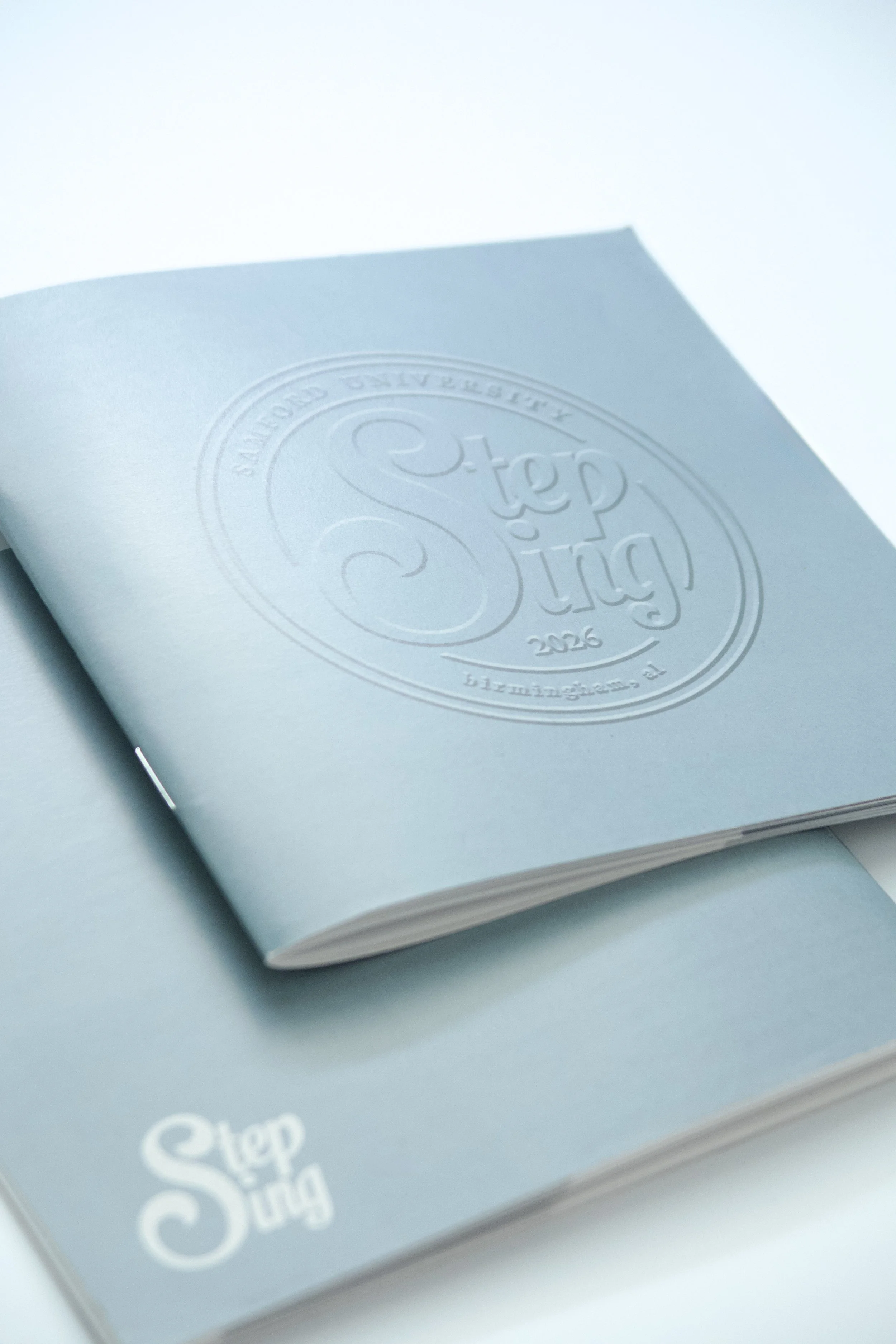Close-up of a light blue notebook with a circular embossed logo on the cover that reads 'Samford University Step Sing 2026'.