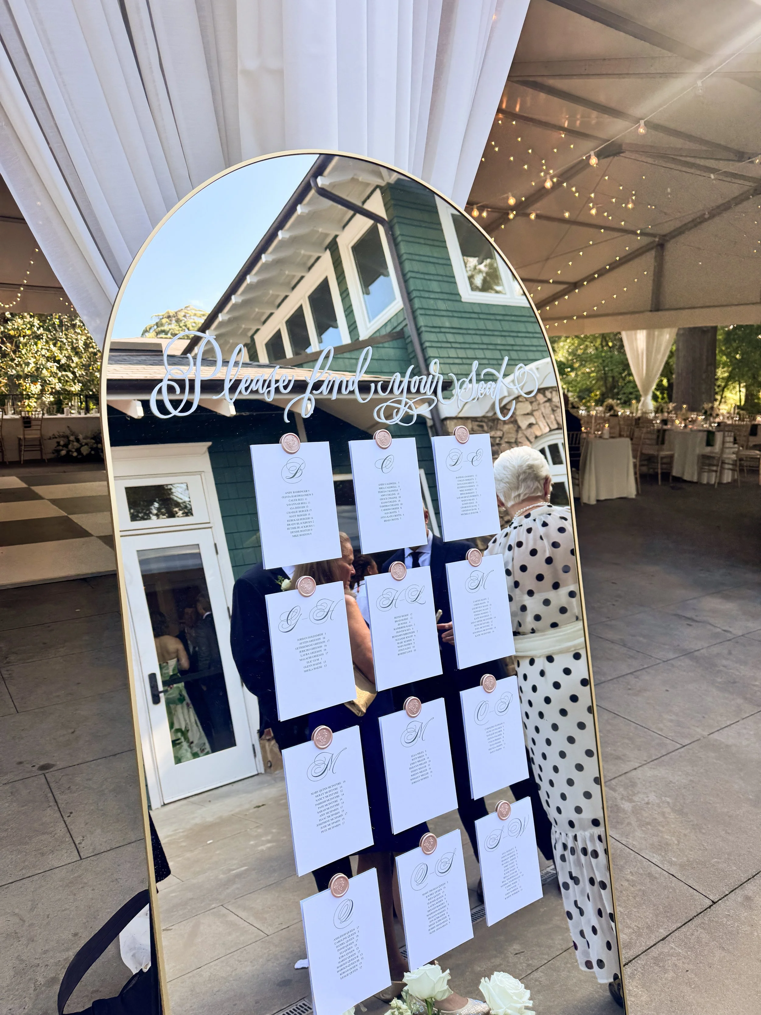 A mirror with the words 'Please Find Your Seat' written on it, displaying a seating chart for an event, and reflecting people dressed in formal attire inside a decorated tent with fairy lights.