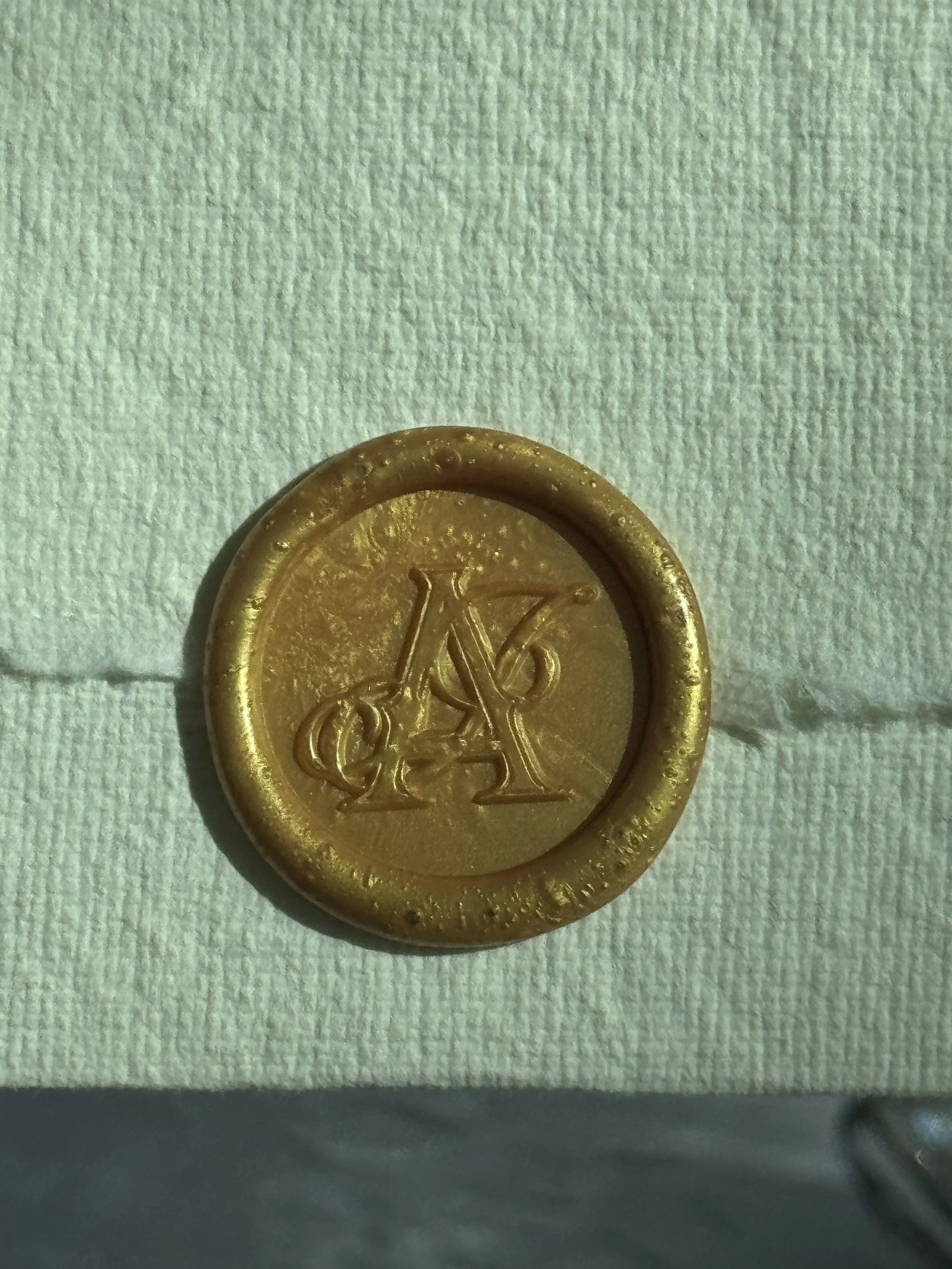 Close-up of a gold-colored wax seal with the duogram of the letters 'A' and 'J' on a white textured card.
