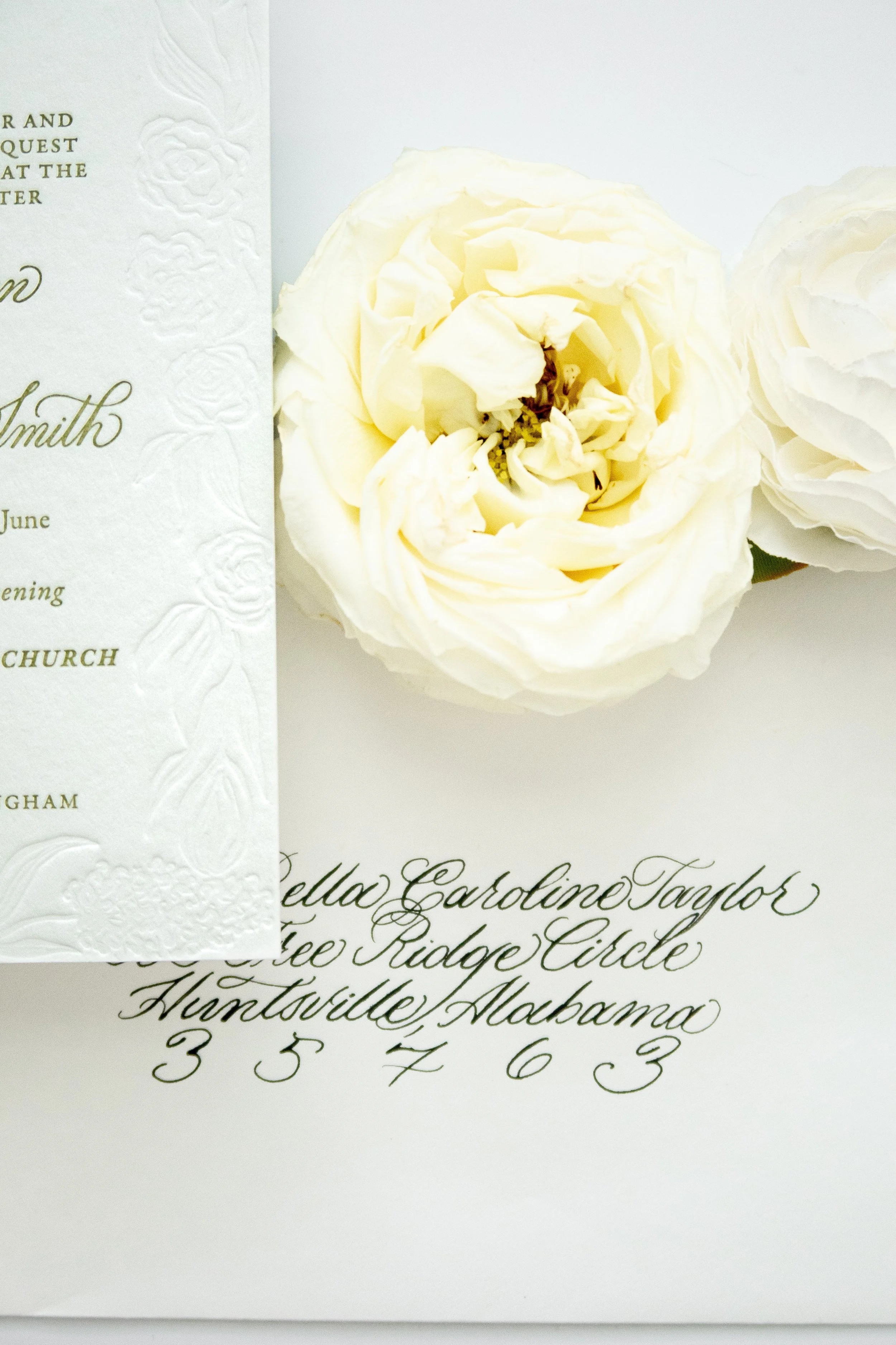 Close-up of an elegant wedding invitation card with embossed floral design on the left side and gold scripted text on the right side, featuring a white flower decoration near the top.