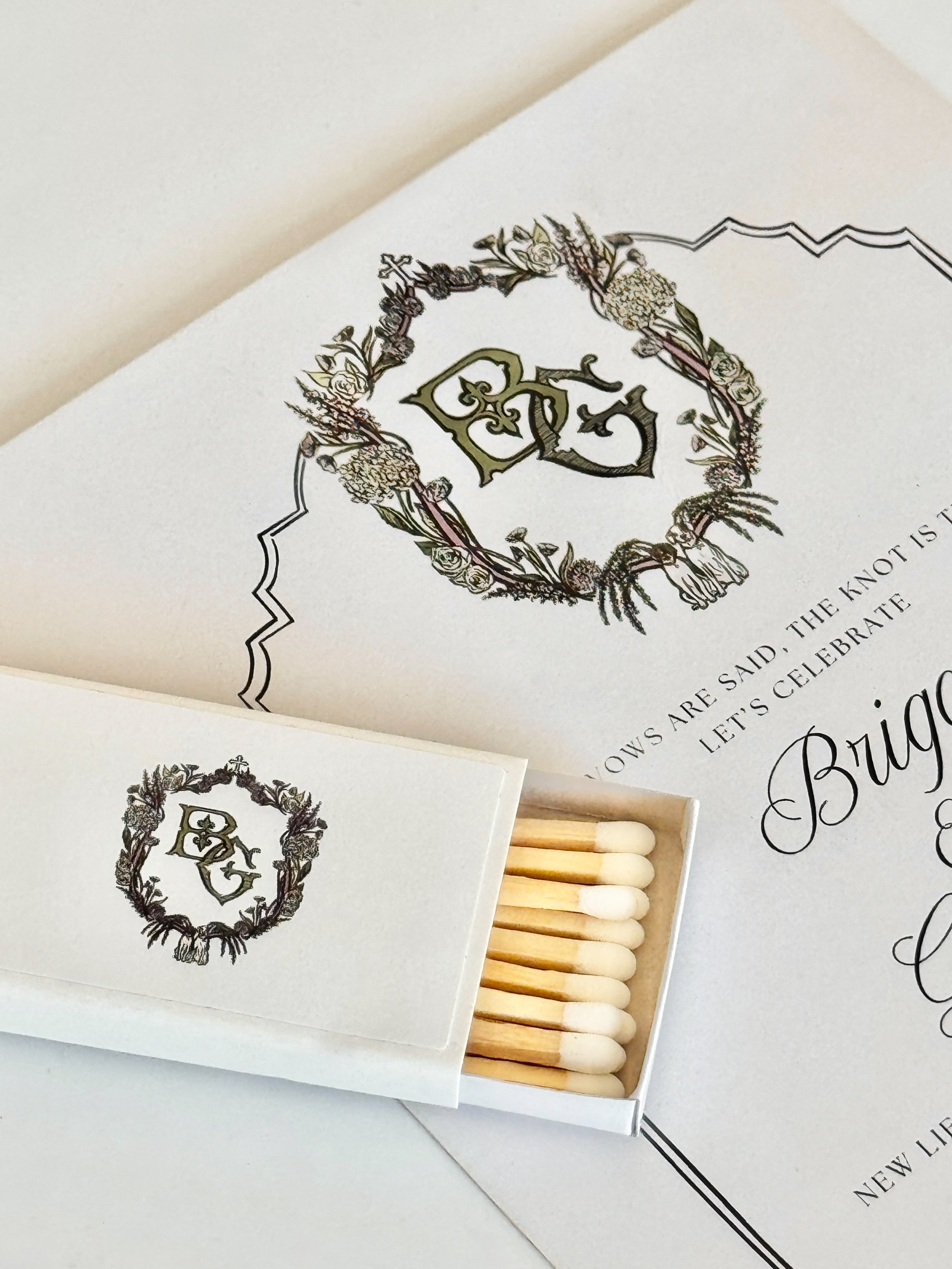 Close-up of wedding stationery with a decorative crest floral design, accompanied by a pack of matchsticks.