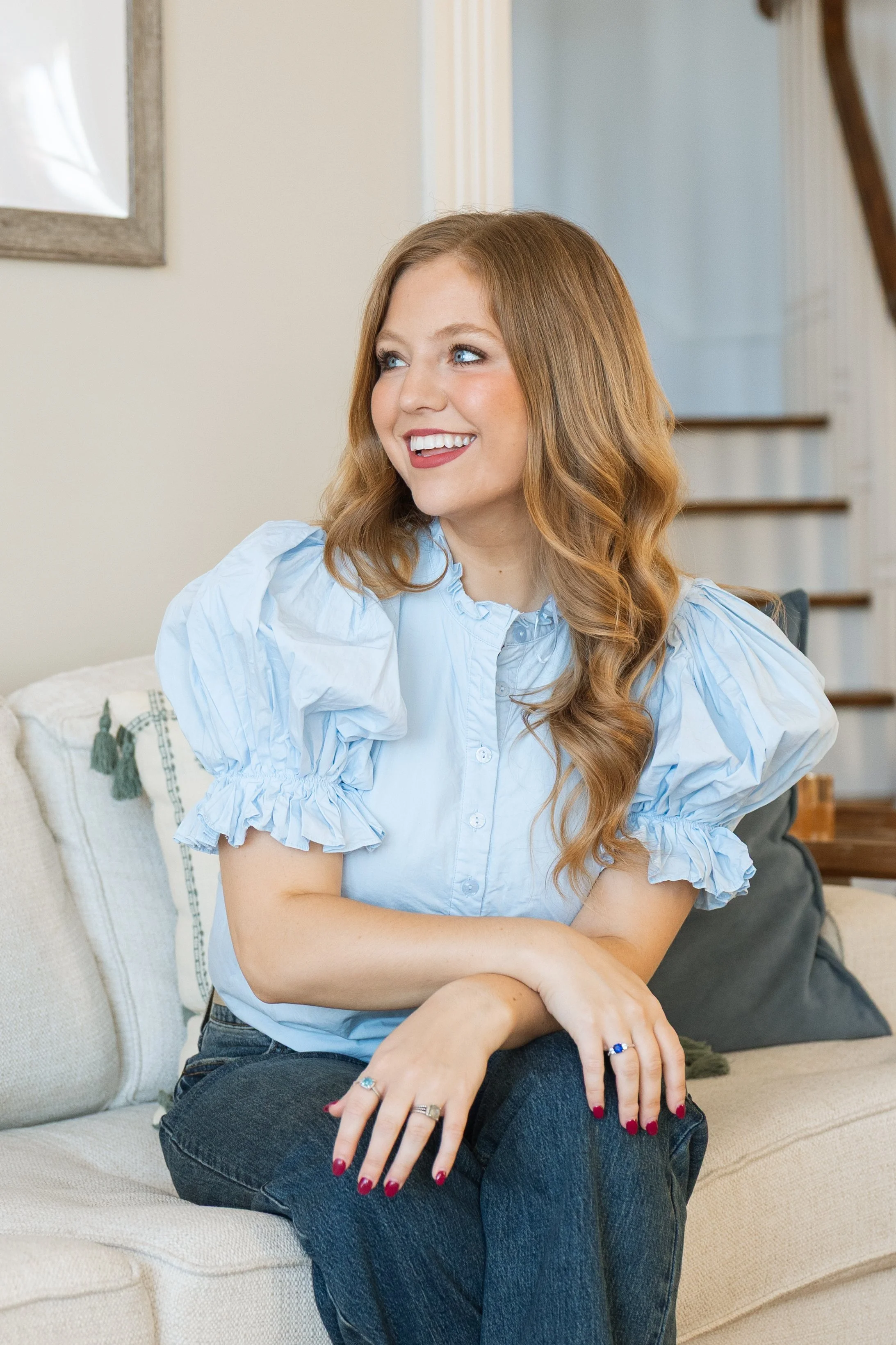 A woman with wavy blonde hair and blue eyes sitting on a beige couch in a living room, wearing a light blue blouse with ruffled sleeves and dark jeans, smiling and looking off to the side.