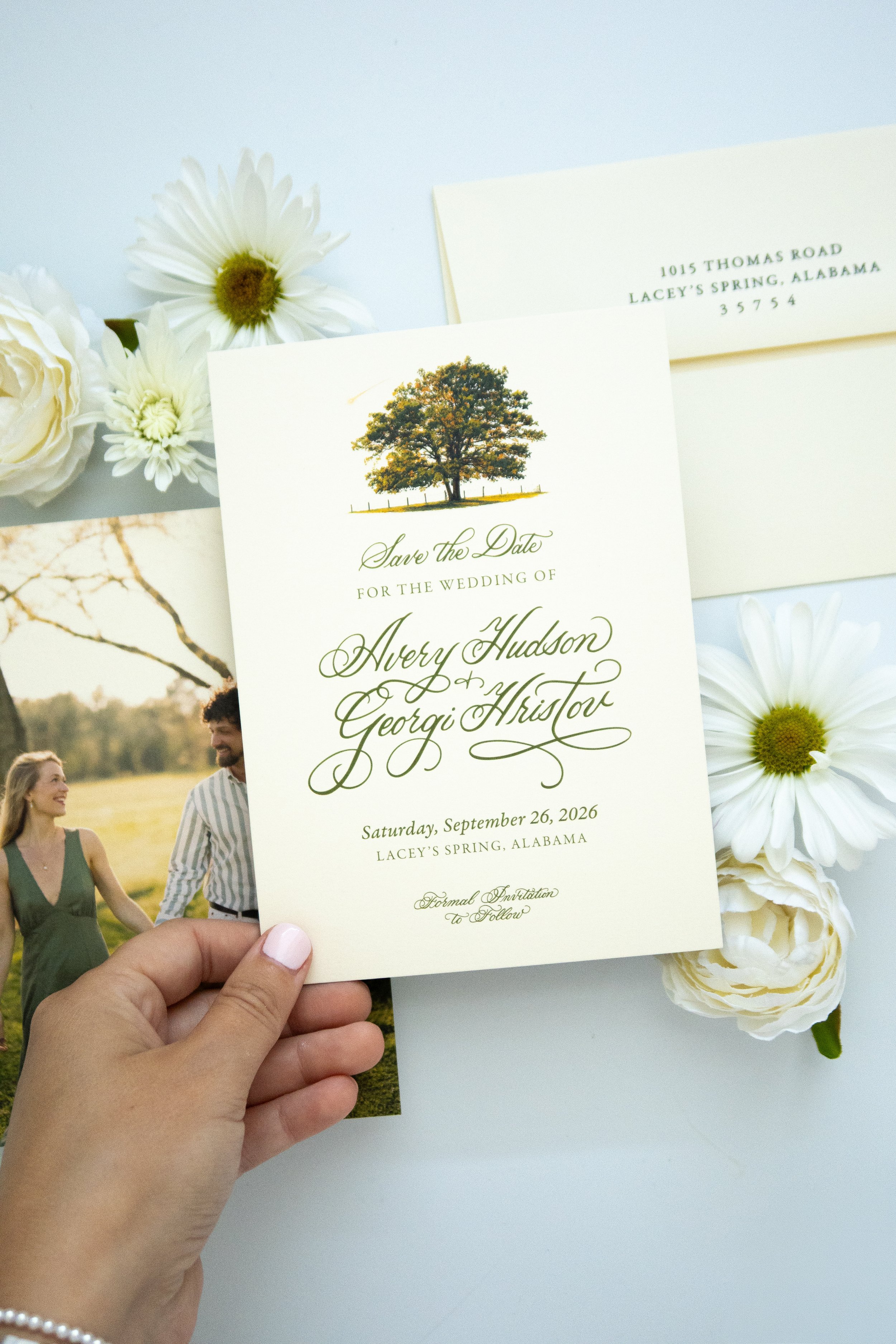 A hand holding a wedding save-the-date card with a tree illustration and elegant cursive text in front of a floral backdrop and outdoor scenery.