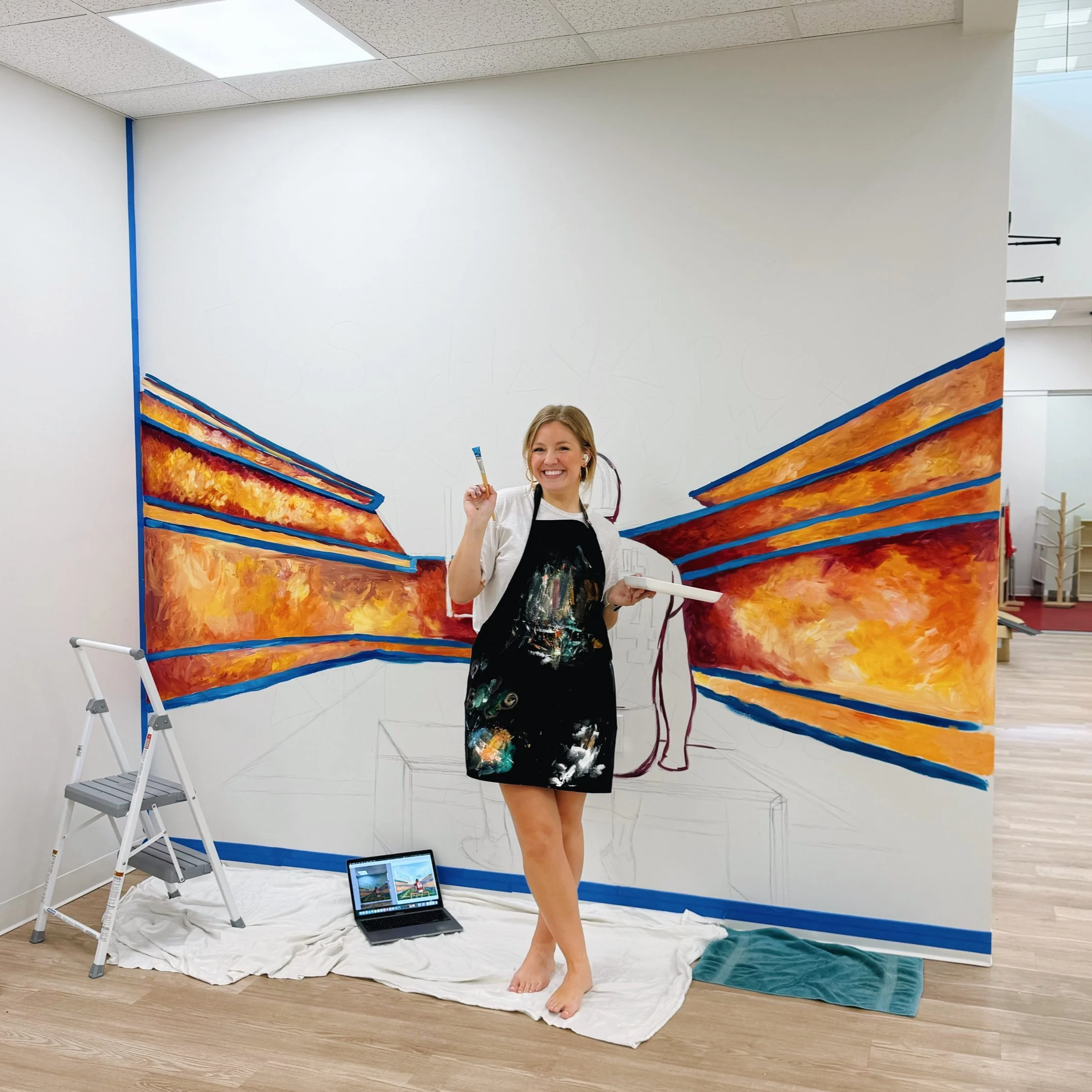A woman with paint on her clothes smiling in front of a large, colorful mural she is painting on a wall inside a room. The mural features vibrant, abstract, orange, yellow, and blue strokes. The woman is holding a paintbrush and standing on a white sheet on the floor, with a laptop showing the painting process nearby.