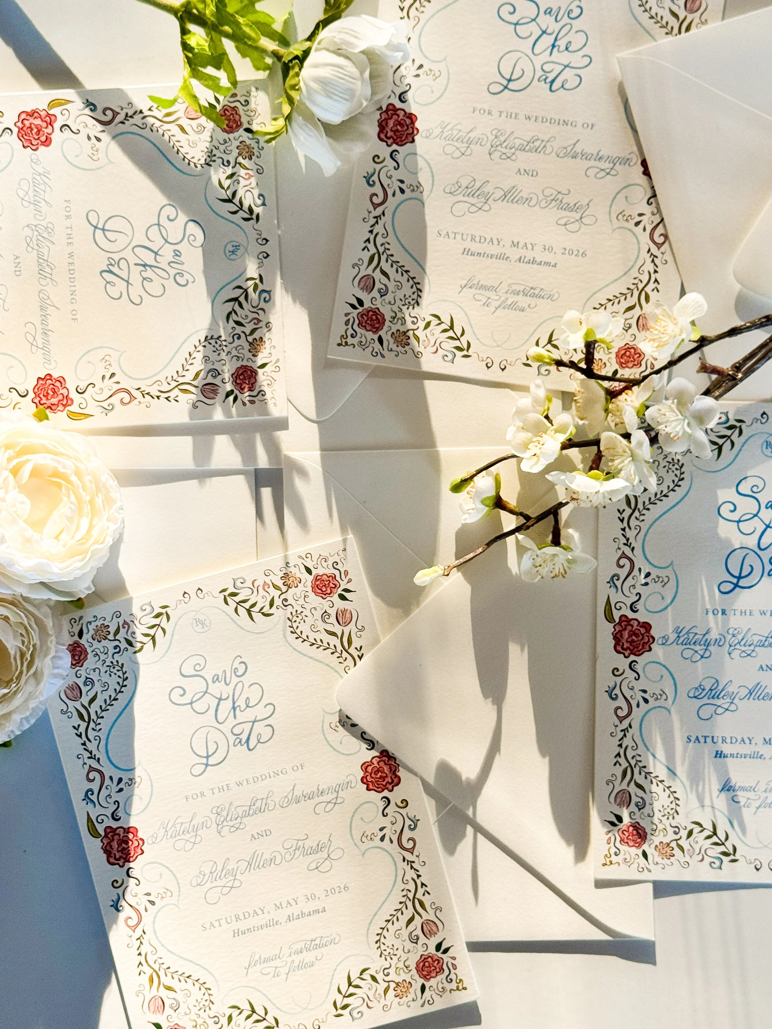 A wedding invitation with floral designs, placed among white flowers and a branch with blossoms. There are also other similar cards and an envelope visible.