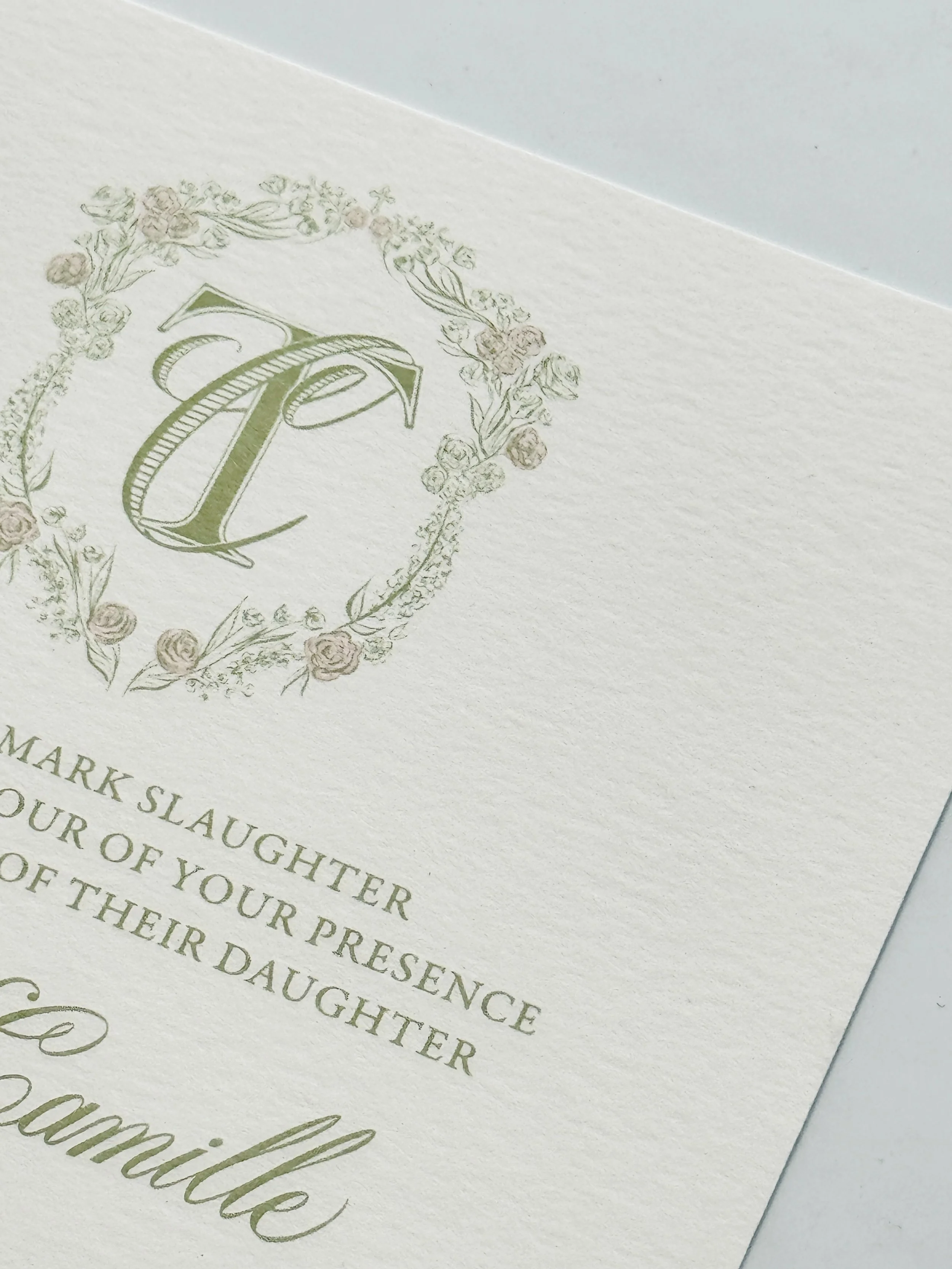 Close-up of a wedding invitation featuring a floral wreath with a large duogram of 'T' & 'C' in the center.