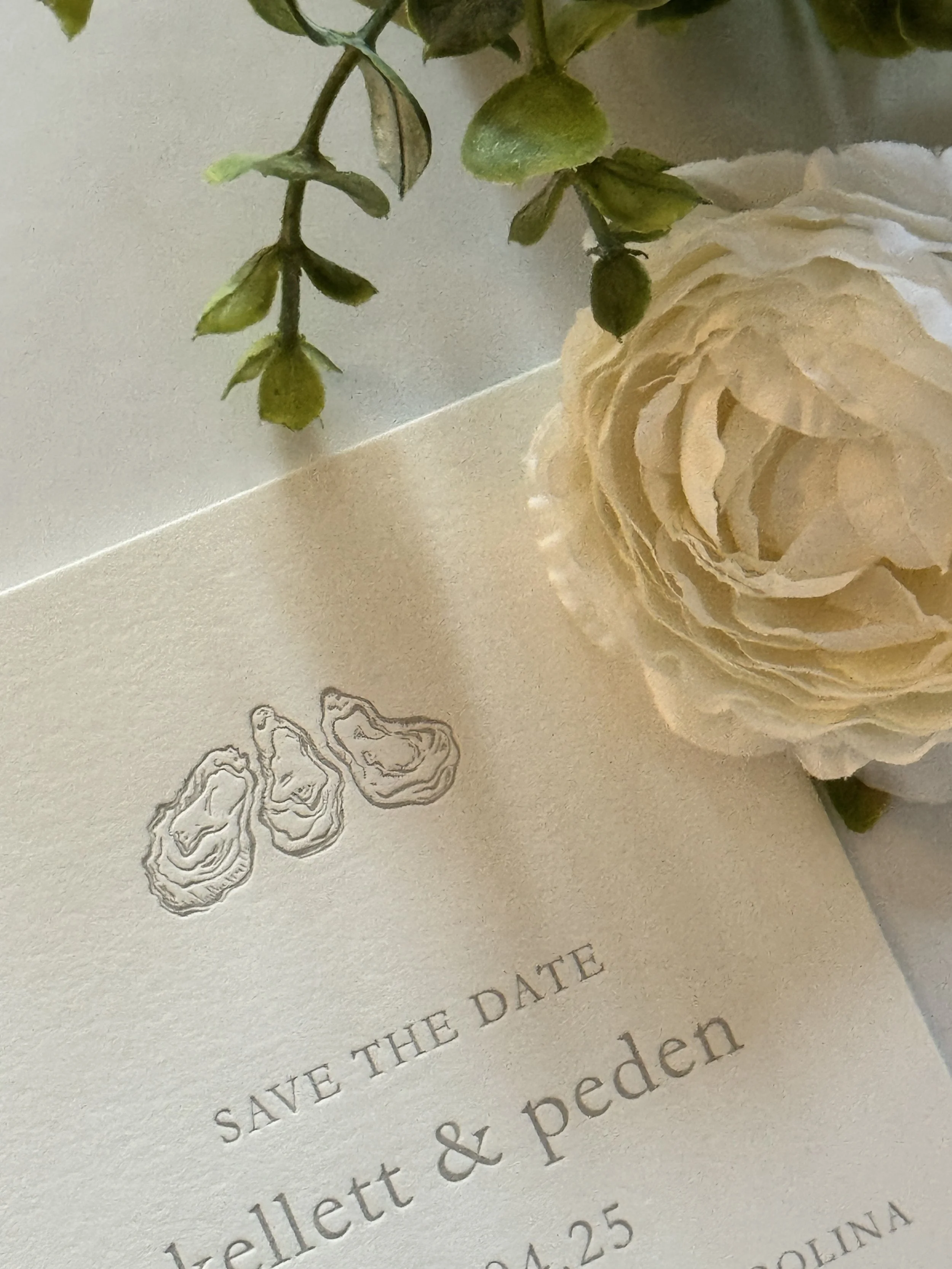 A close-up of a wedding invitation with a white rose and green leaves decoration, a small illustration of layered oysters, and text with the date of the wedding.
