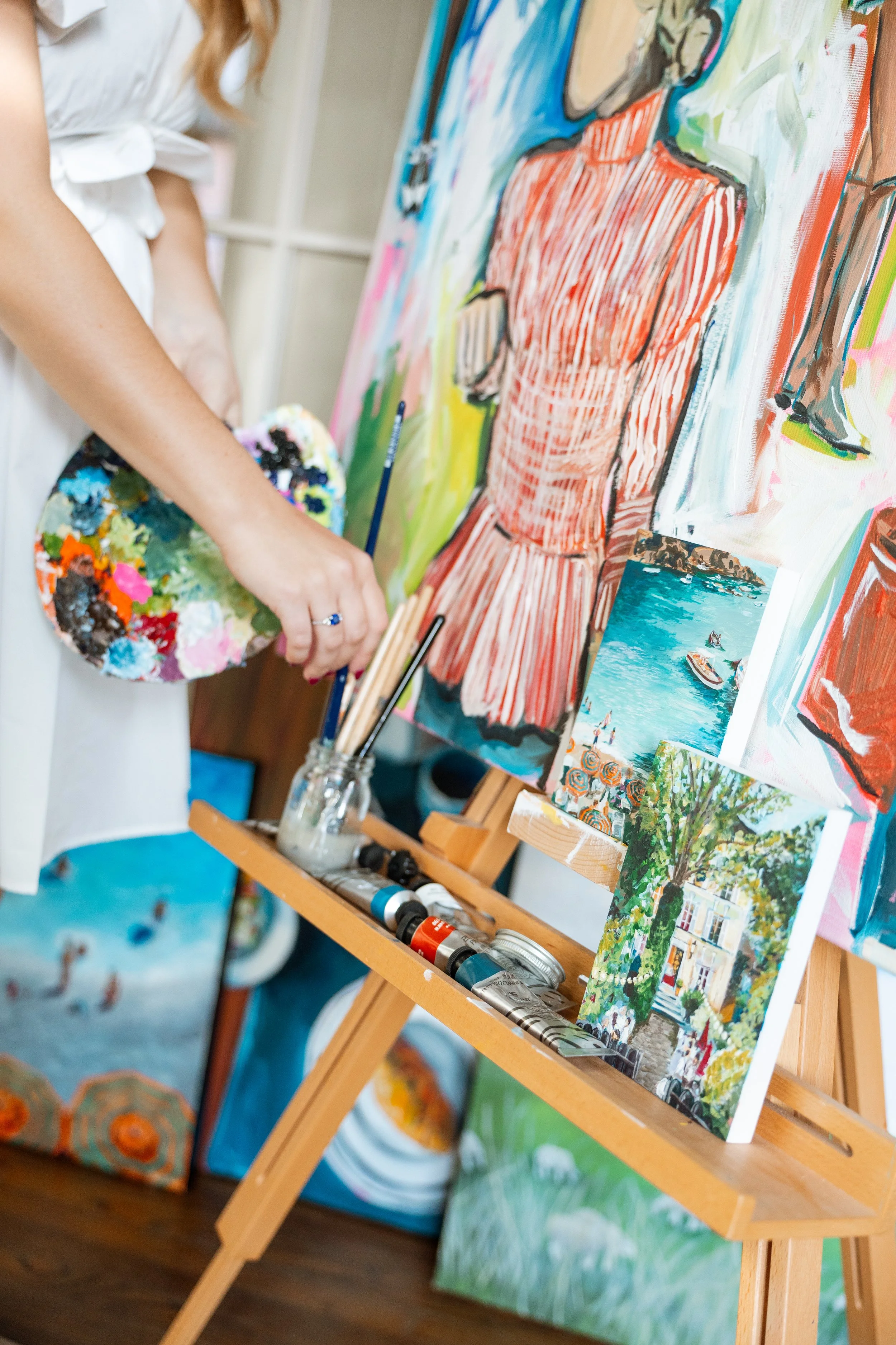 A woman in a white shirt holding a painter's palette full of colorful paints, standing in front of a wall displaying several painted landscapes and scenes. The scene suggests she is painting or viewing her artwork in an art studio.