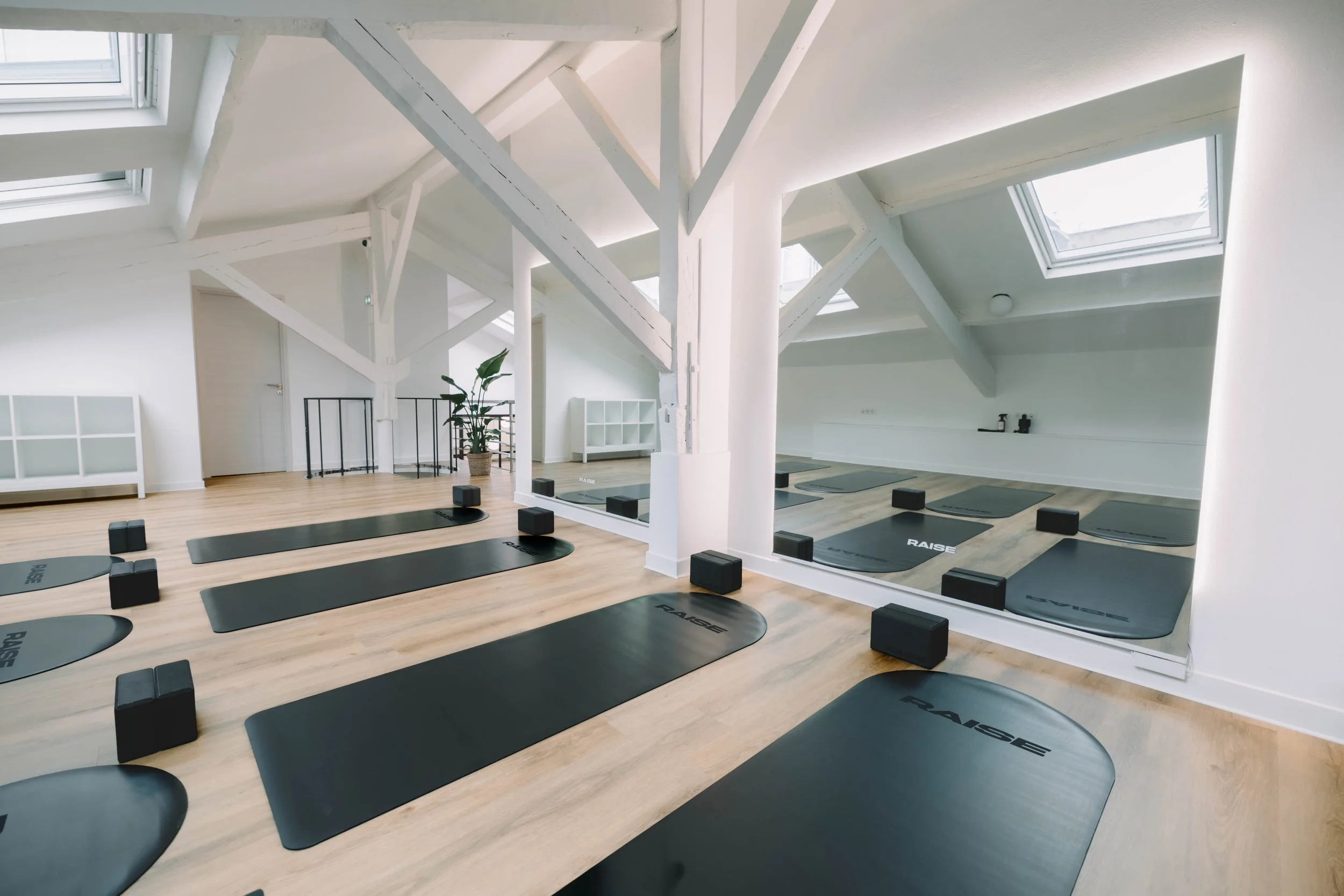 Salle de yoga lumineuse avec tapis noirs et blocs de soutien, grande fenêtre de toit, décoration minimaliste, miroir mural et plante verte.