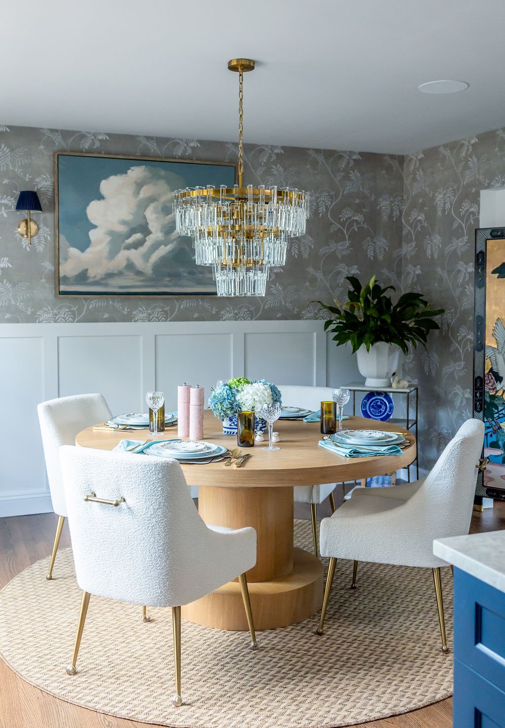 Elegant dining room with a round wooden table set for four, decorated with floral centerpiece, pink and amber glasses, and plate settings. Surrounded by cream-colored upholstered chairs with gold legs. The room features a chandelier with crystal accents, a large piece of sky-themed artwork, a green leafy plant in a white vase, and patterned gray wallpaper.
