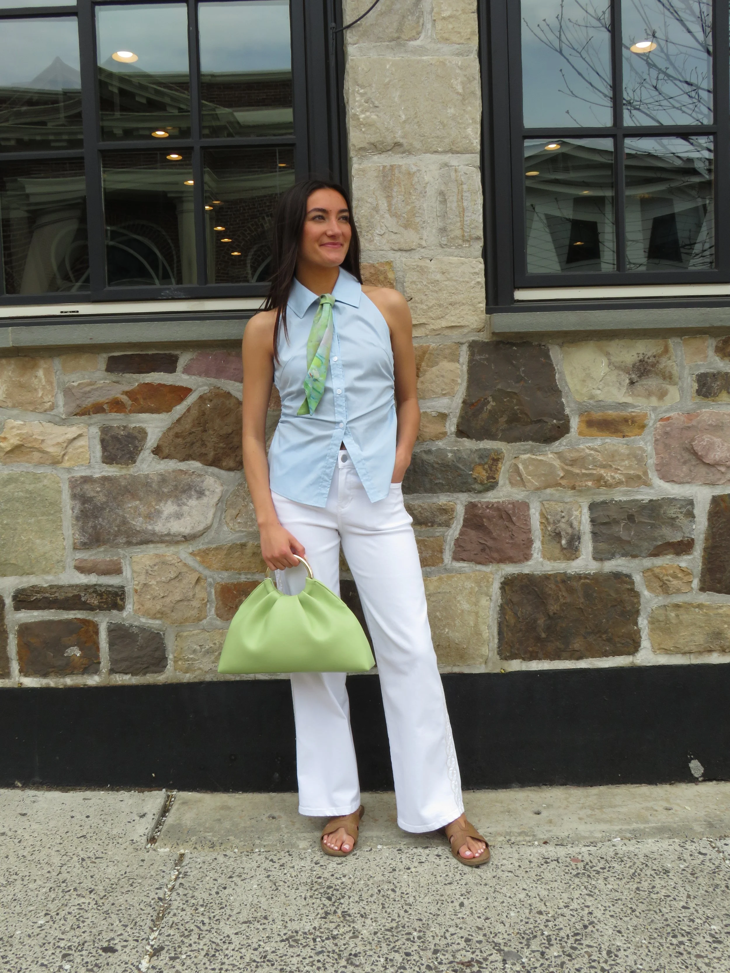 A woman with long dark hair wearing a light blue sleeveless button-down shirt, white wide-leg pants, tan sandals, and a light green handbag, standing outside against a stone wall with windows reflecting the cloudy sky and nearby buildings.