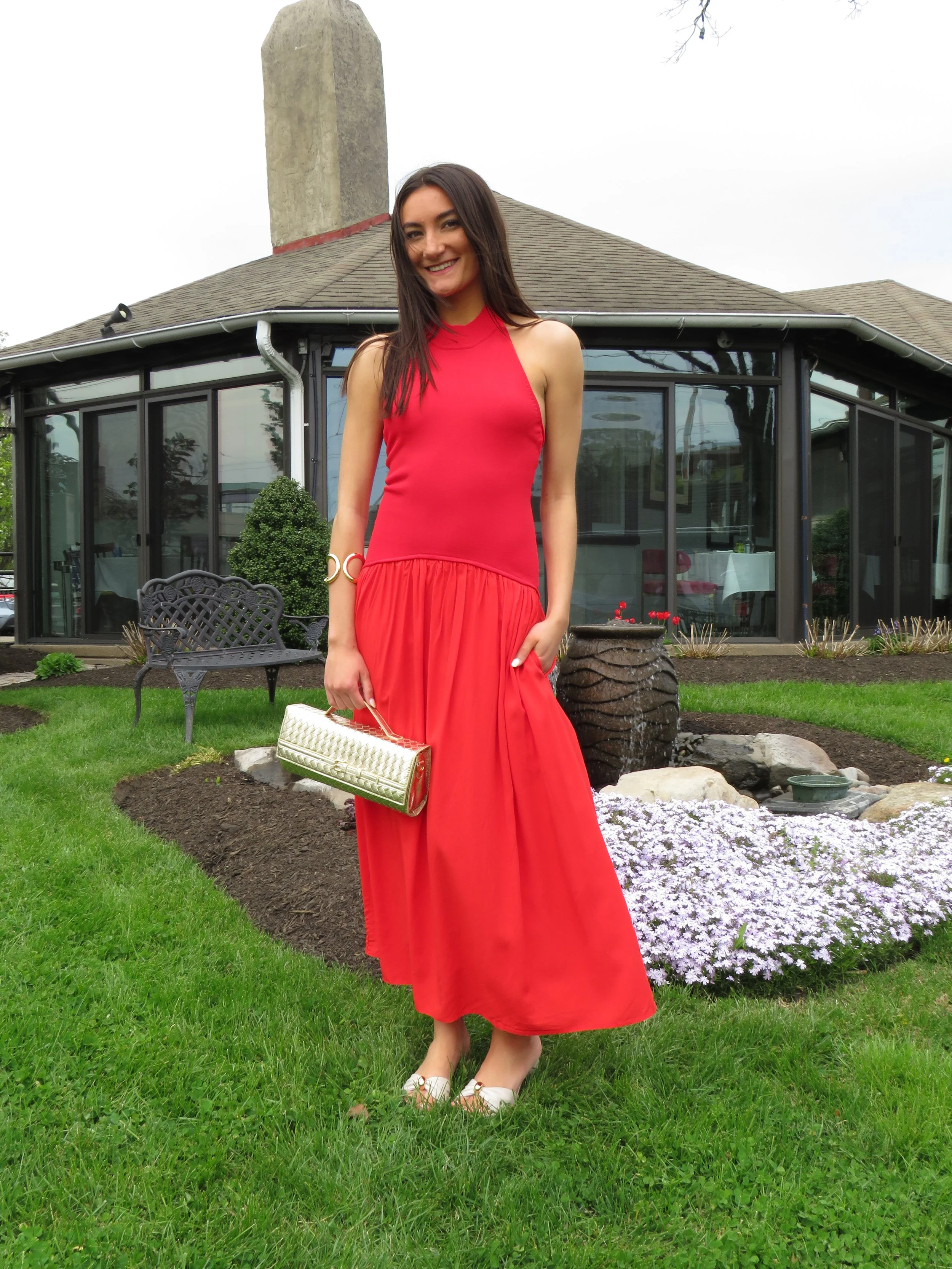 A woman in a red dress holding a beige clutch purse poses in a garden with a background of a house with glass windows and a chimney. The garden features purple flowers and a small fountain.