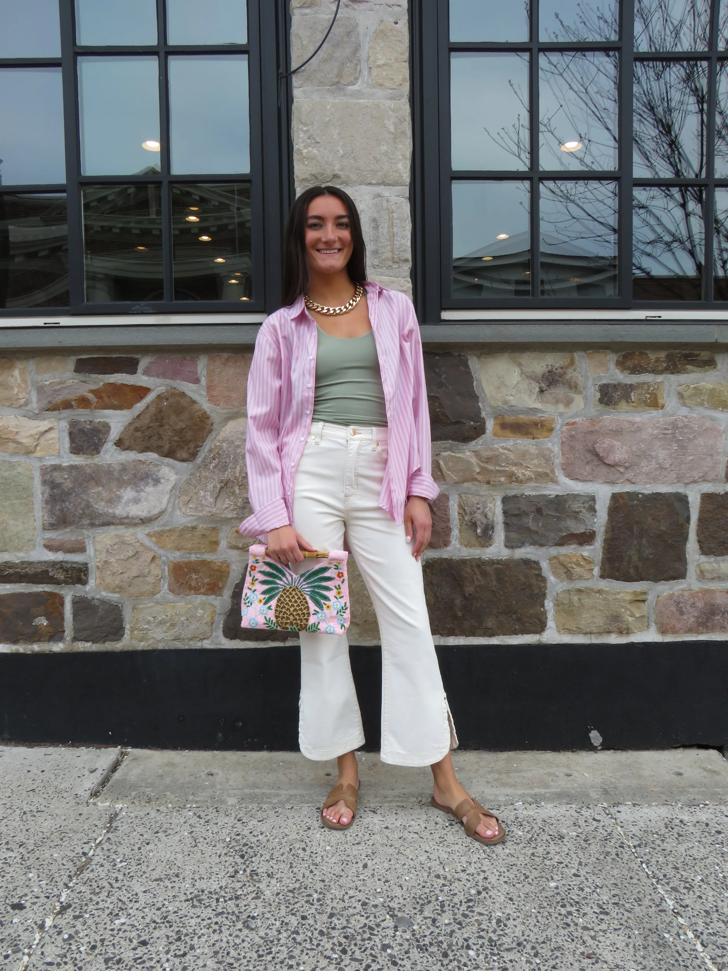 A woman standing in front of a stone wall with large windows, wearing a pink and white striped shirt, white pants, tan slide sandals, a gold chain necklace, and holding a colorful pineapple-themed tote bag.