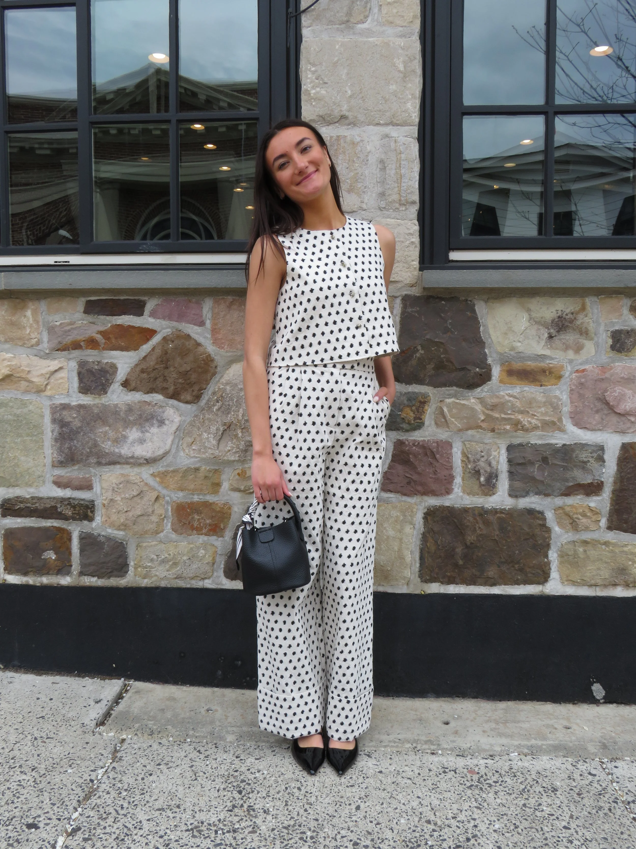 A young woman with dark hair and light skin, smiling, in a polka dot sleeveless top and matching polka dot pants, holding a black handbag with a striped scarf, standing in front of a stone building with large window panes.