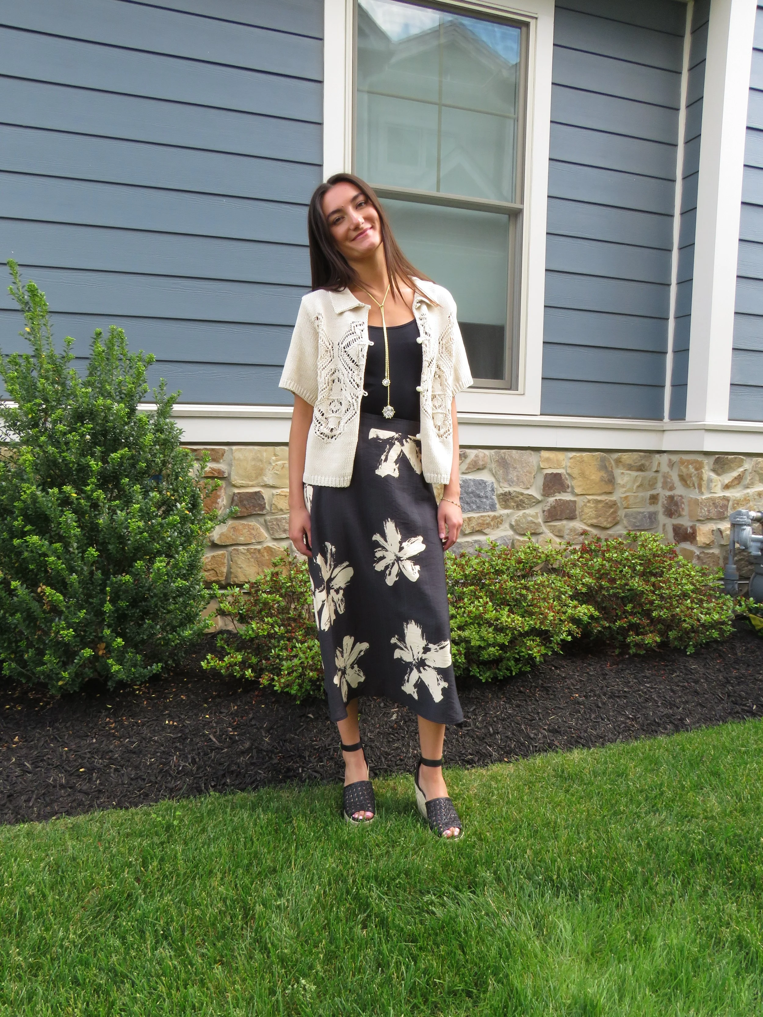 A woman standing outdoors on a green lawn in front of a house with blue siding and a stone foundation. She is smiling, wearing a black dress with a floral pattern, a cream lace cardigan, and black high-heeled sandals.