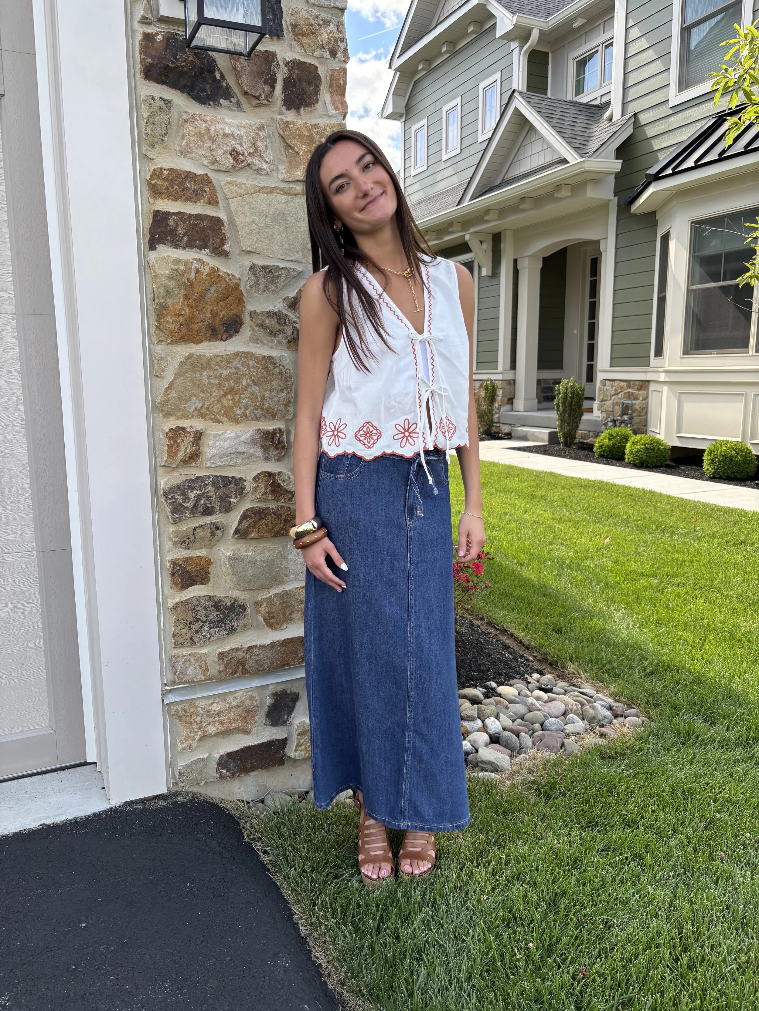 A woman standing outside in front of a house, wearing a white sleeveless top with red embroidery, a long denim skirt, and brown high-heeled sandals, smiling at the camera.