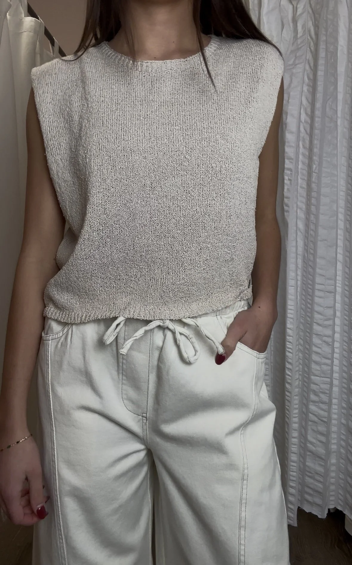 Woman wearing a sleeveless knitted beige top and white drawstring pants, with her right hand in her pocket.