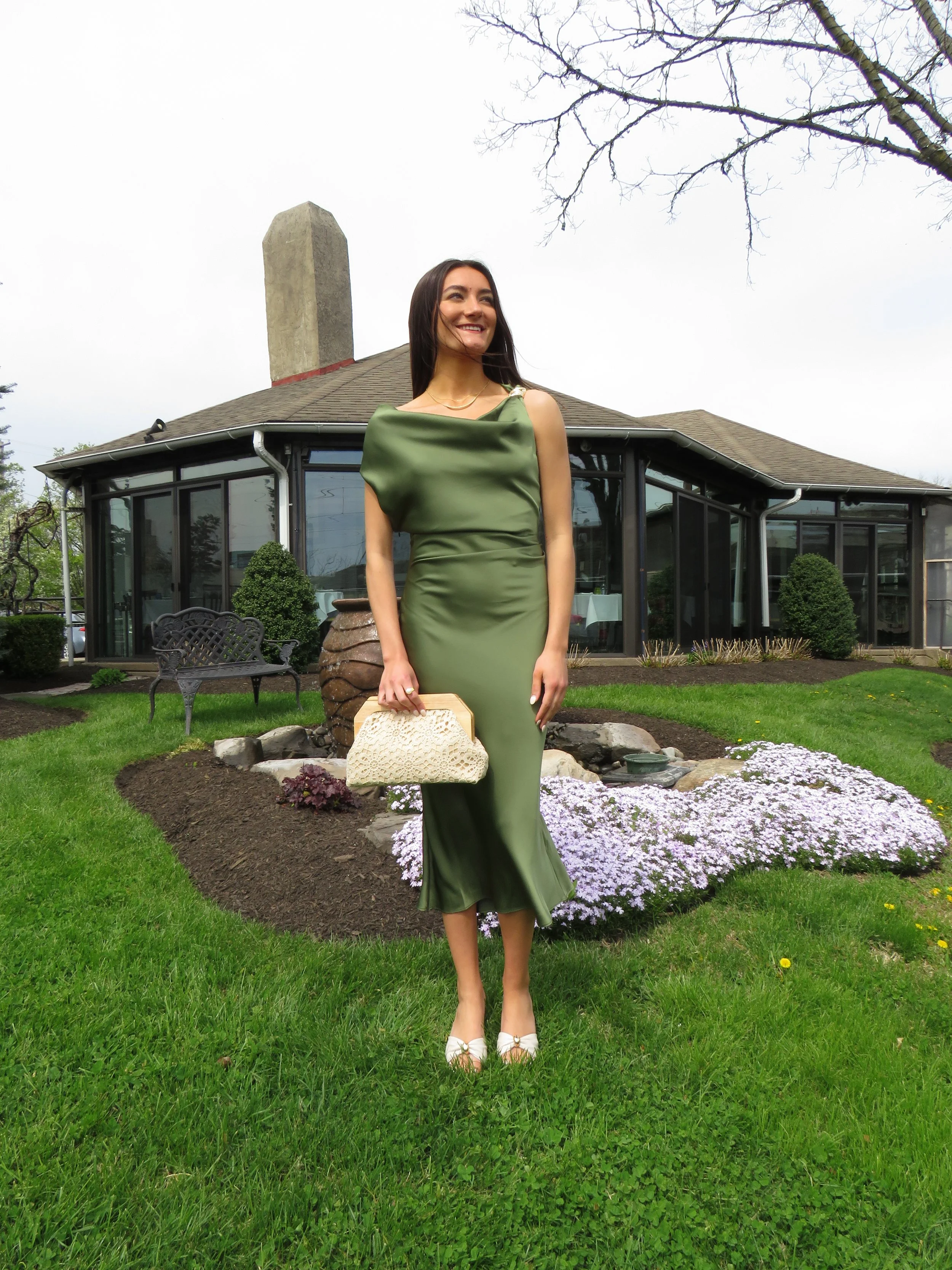 A woman in a green satin dress standing on a garden lawn, holding a cream-colored handbag, with purple and white flowers and a house with large windows behind her.