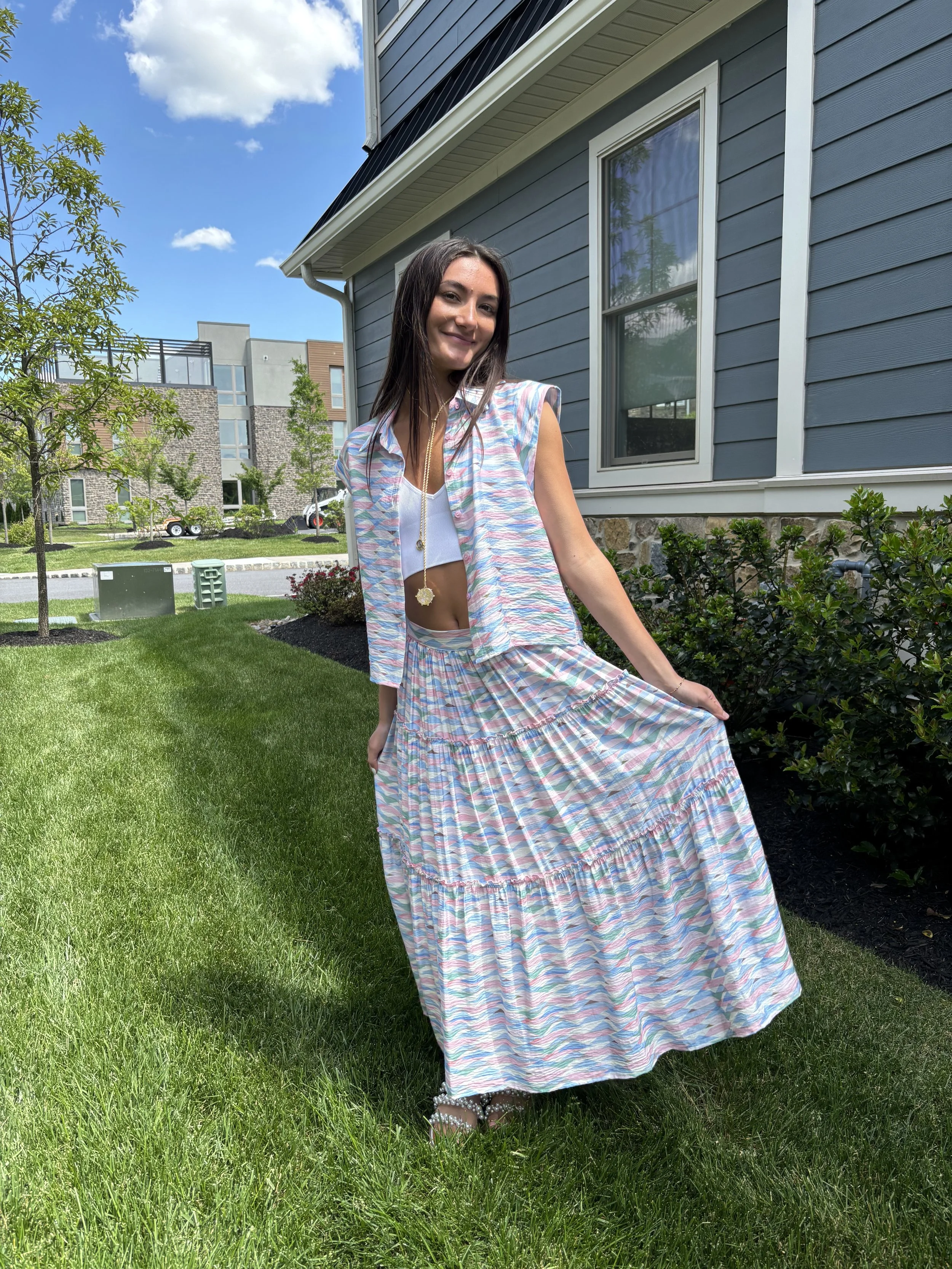 A young woman in a colorful, pastel-patterned dress posing on a well-manicured lawn in front of a gray house with white trim. She is smiling, looking at the camera, and holding up her long skirt with one hand. The scene is sunny with a blue sky and s